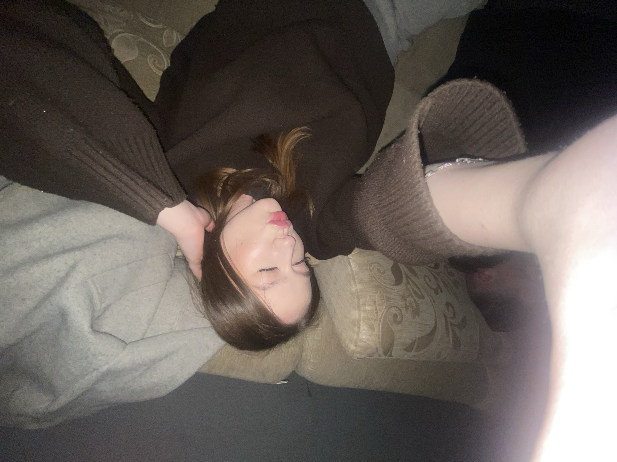 A close-up, low-angle selfie captures a young person lying down indoors, likely sleeping or resting. The subject, with long brown hair, is wearing a dark brown knitted sweater or top. Their eyes are closed and lips are slightly parted, suggesting a state of relaxation or sleep. One hand is positioned near their head, while the other arm, adorned with a silver-colored bracelet or chain, is extended towards the camera, indicating it is likely holding the device that took the photo.
The person is lying on a light-colored couch or armchair featuring an elaborate, possibly floral or scroll-patterned upholstery. A light grey, possibly quilted, garment or blanket is visible on the left side of the frame, partially covering the subject. Another plain grey surface is visible in the lower foreground.
The lighting is dim, suggesting it is evening or night, or a dimly lit room, with a bright flash illuminating the right side of the image. The overall mood is quiet and intimate. There is no visible text in the image. The scene is set in Chaikovsky, Russia.
