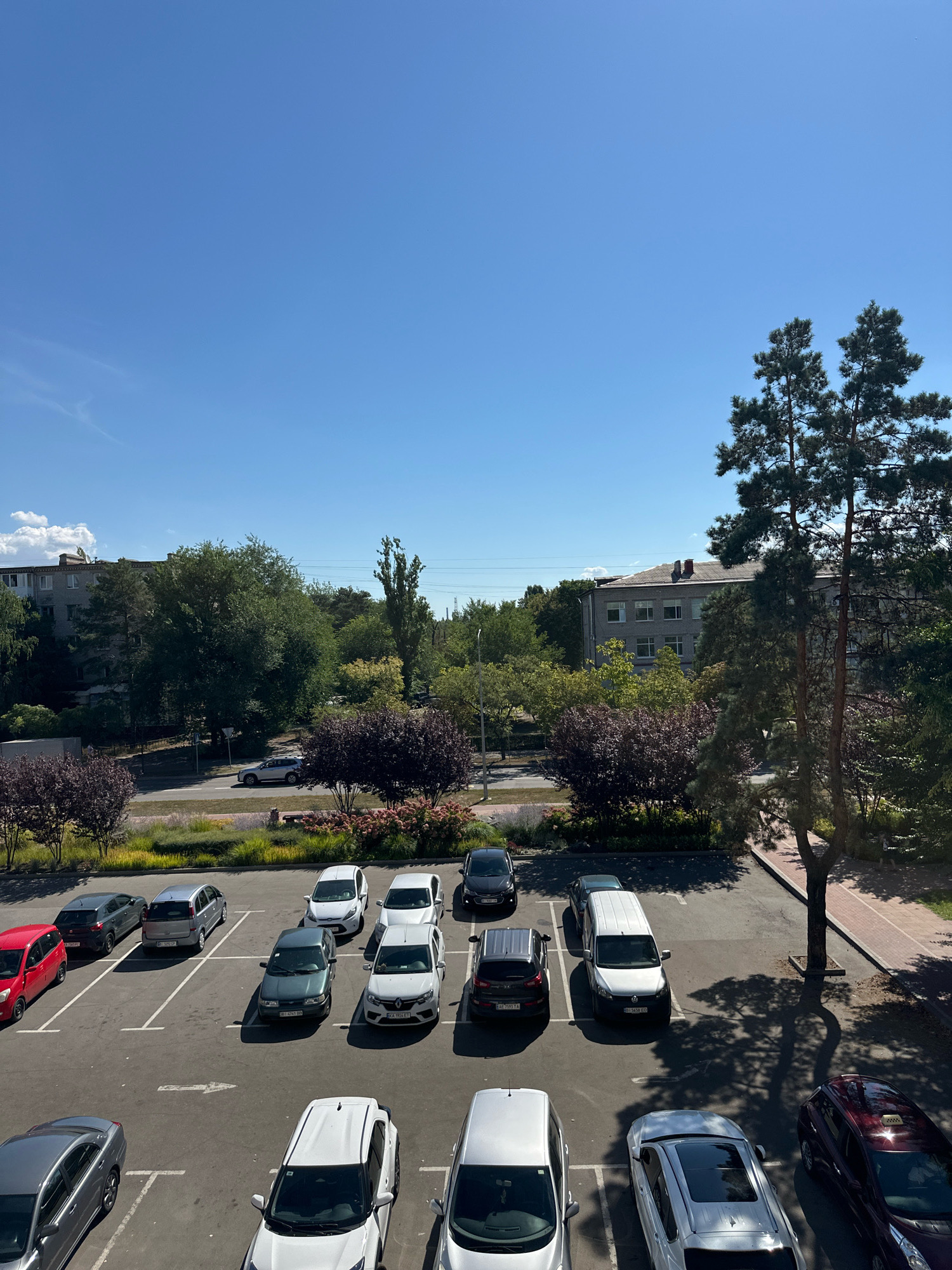 The image shows a parking lot in the city of Horishni Plavni, Ukraine, on a sunny day. The sky is clear and blue, with a few wispy clouds. In the foreground, there are several cars parked in marked spots. These cars include a red hatchback, a dark gray sedan, a silver minivan, a dark green sedan, a white compact car, a white hatchback, and a dark gray SUV. Further back, there are more vehicles visible in a street and another parking area.
Behind the parking lot, there is a strip of landscaping with trees and bushes. Some of the bushes have deep purple foliage, while others have pink flowers. Beyond the landscaping, there are buildings, including a multi-story apartment building and what appears to be another residential or institutional building. Tall evergreen trees and other deciduous trees are interspersed throughout the scene, casting shadows on the ground. A paved walkway or sidewalk runs alongside the parking lot on the right side of the image. The overall impression is of a typical urban residential or commercial area during daylight hours.