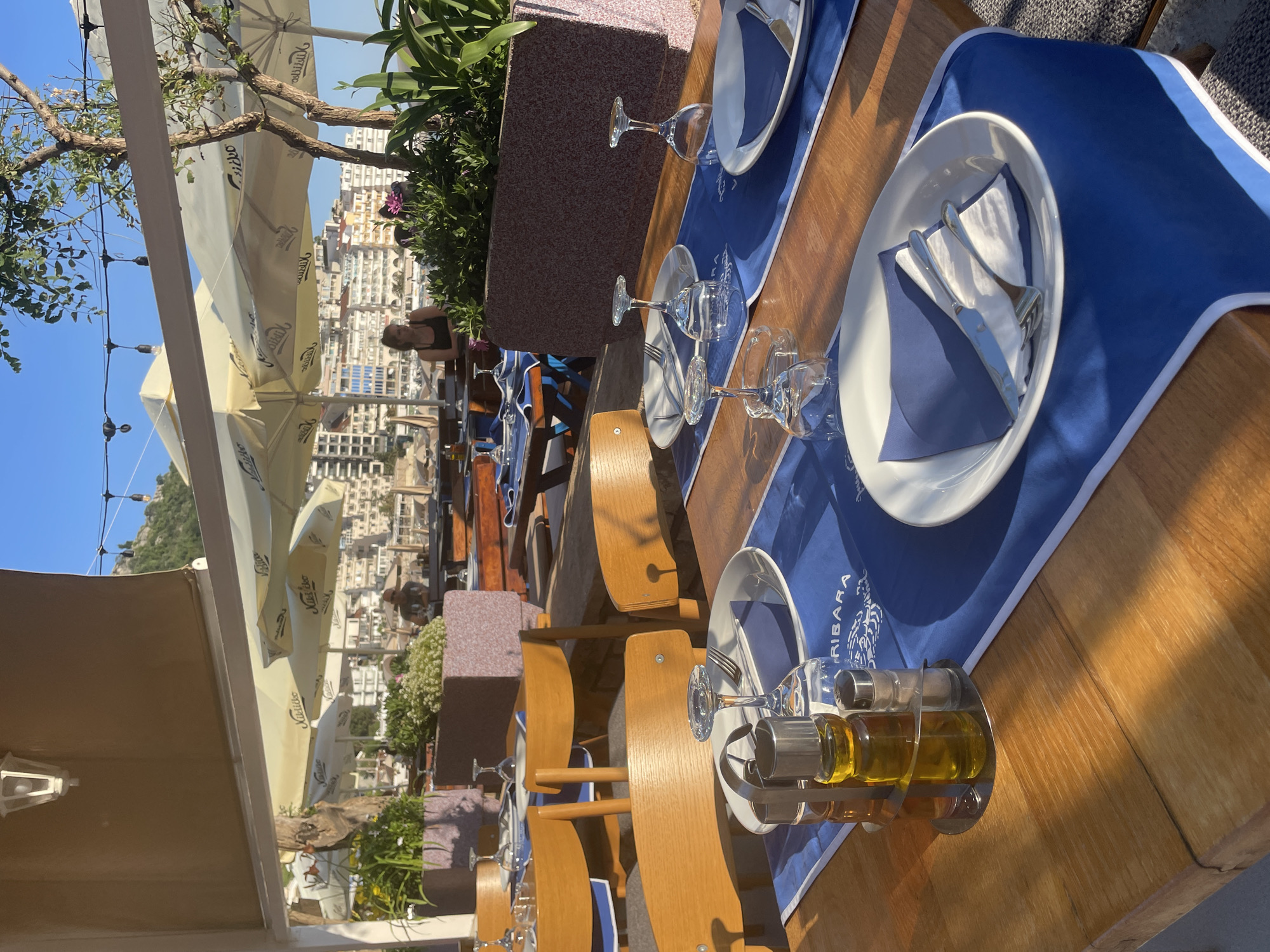 The image shows an outdoor restaurant setting in Rafailovići, Montenegro. The tables are set with blue placemats, white plates, silverware, wine glasses, and oil/vinegar dispensers. Wooden chairs are arranged around the tables. In the background, there's a view of the city buildings and a glimpse of the sky. There are parasols providing shade. A person is sitting at one of the tables in the distance. The restaurant seems to be on a high terrace or balcony.