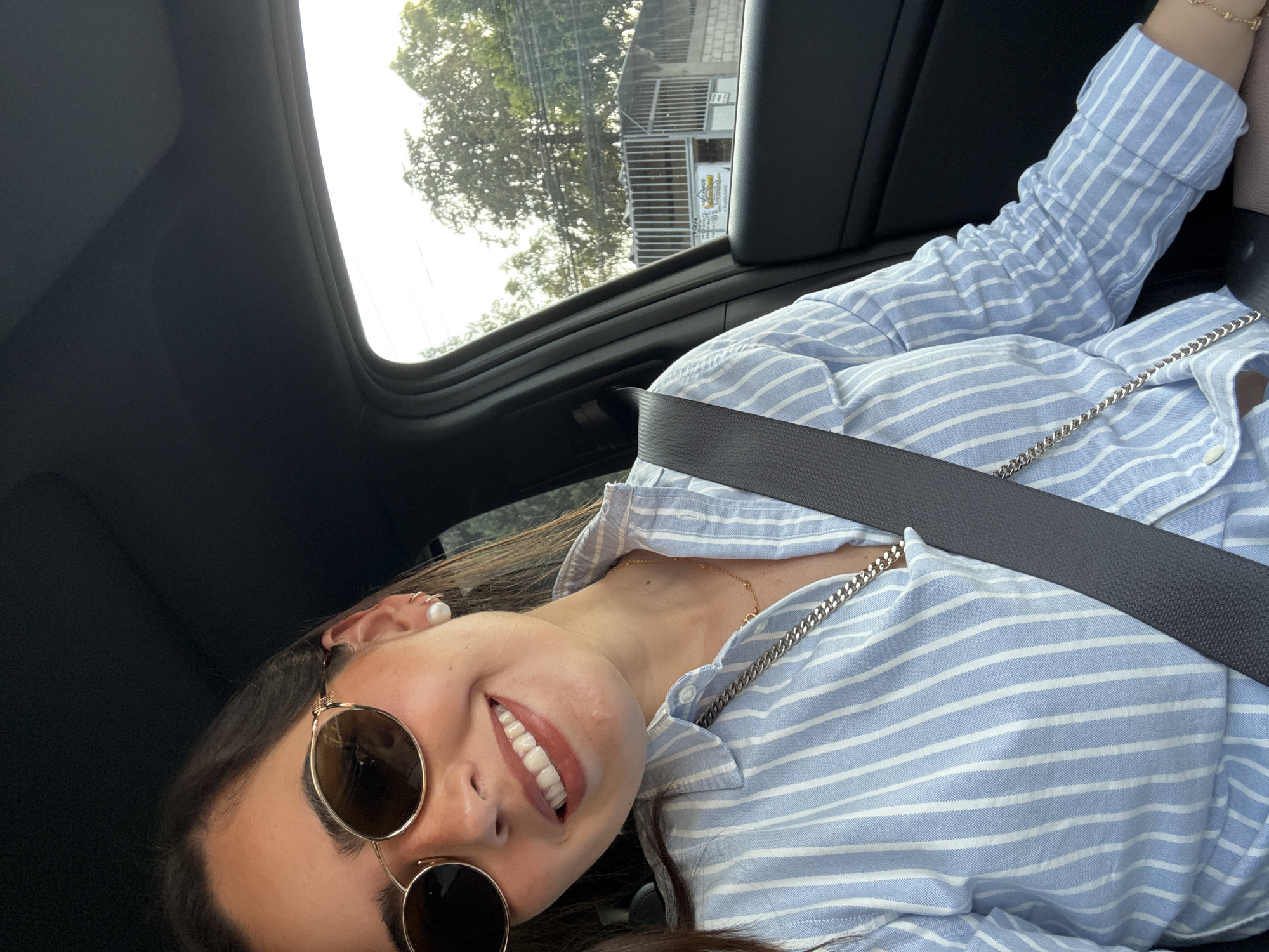 A woman is seated in a vehicle, likely a car, wearing a blue and white striped shirt, sunglasses, and a seatbelt. She is smiling. Outside the car window, a tree and a building can be seen. The location is likely the city of Puebla, Mexico, based on the style of the buildings and the foliage.