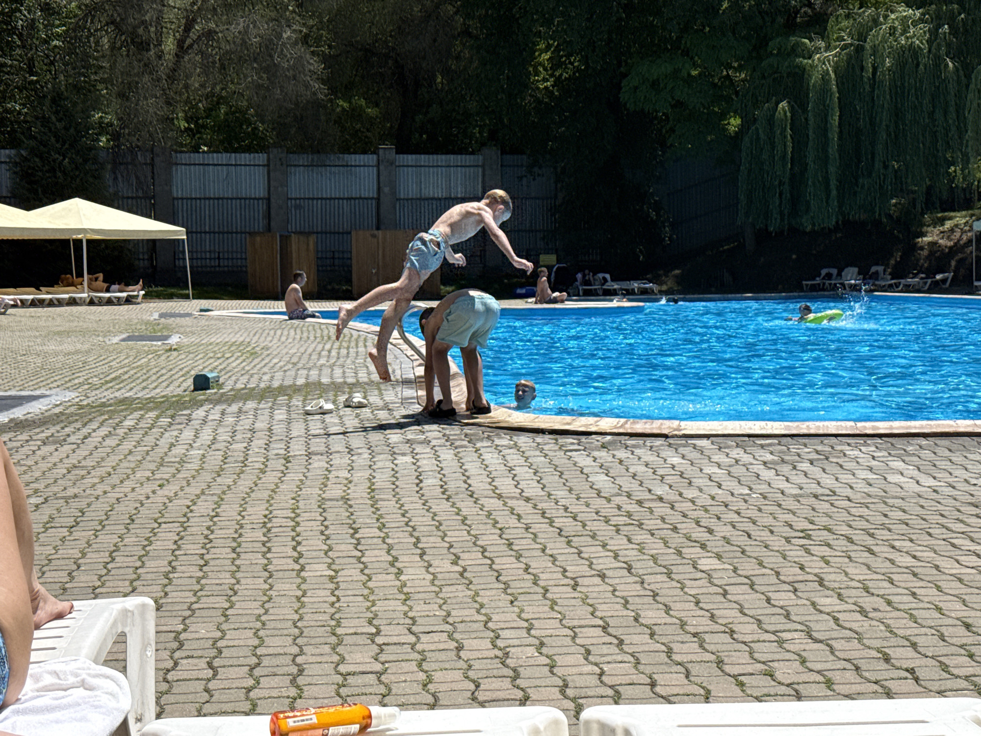 The image depicts a bright, sunny day at an outdoor swimming pool, likely a public or private facility, situated within a city park or recreational area. The pool itself is large and filled with clear blue water, with ripples indicating activity. There are several people present. In the foreground, a young boy in blue swim trunks is captured mid-jump, poised to enter the pool. Another person, possibly an adult, in light blue shorts, is bending down near the edge of the pool, appearing to be watching the jumper or waiting to enter. In the background, several other individuals are visible, some lounging on chairs under umbrellas, and others in the water, including one person on a yellow float. The poolside area is paved with interlocking brick pavers, and lined with lounge chairs. Beyond the pool area, trees and greenery suggest a natural, park-like setting, and a chain-link fence is visible. The architecture and ambiance are consistent with many of Almaty's recreational facilities, such as the numerous water parks and public pools found within and around the city, which are popular during the warm summer months.