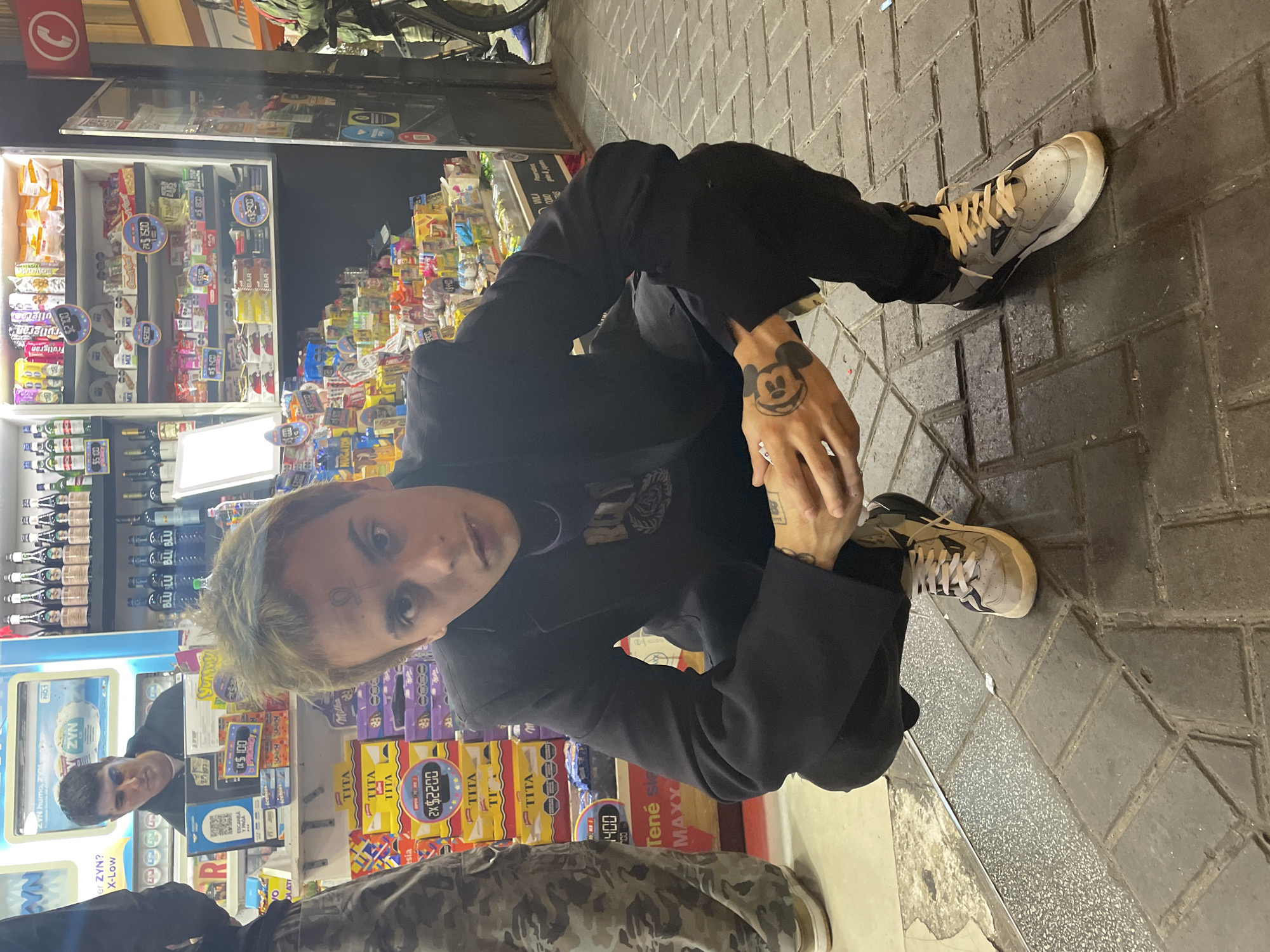 The image shows a person crouching on a sidewalk made of gray, rectangular pavers, likely in Buenos Aires, Argentina, given the Argentinian product branding visible in the background. The individual has light-colored hair, a tattoo under their left eye, and a tattoo of Mickey Mouse on their right hand. They are wearing a black jacket and black pants, with white and gray sneakers. The person is looking directly at the camera. Behind them is a convenience store displaying various packaged goods, including alcohol, candies, and snacks. Price tags are visible on the shelves, written in Argentinian pesos. Another person is partially visible behind the crouching individual, standing inside the store.