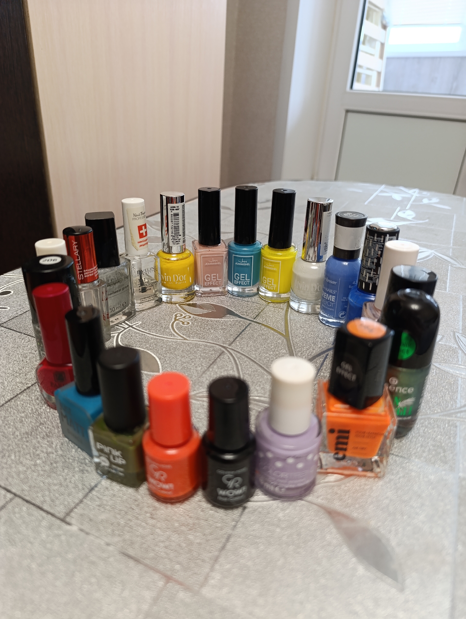 The image shows a collection of nail polish bottles arranged in a circle on a patterned surface, likely a tablecloth. There are approximately 20 bottles of various colors and brands, including Stellary, Nail The Professional, Eveline, Vin D'or, Jeanmishel, Hansen, GR Wow!, Emi, and Essence. The bottles vary in size and shape. The background includes a wall and a doorway or window. The setting appears to be indoors, potentially a home.