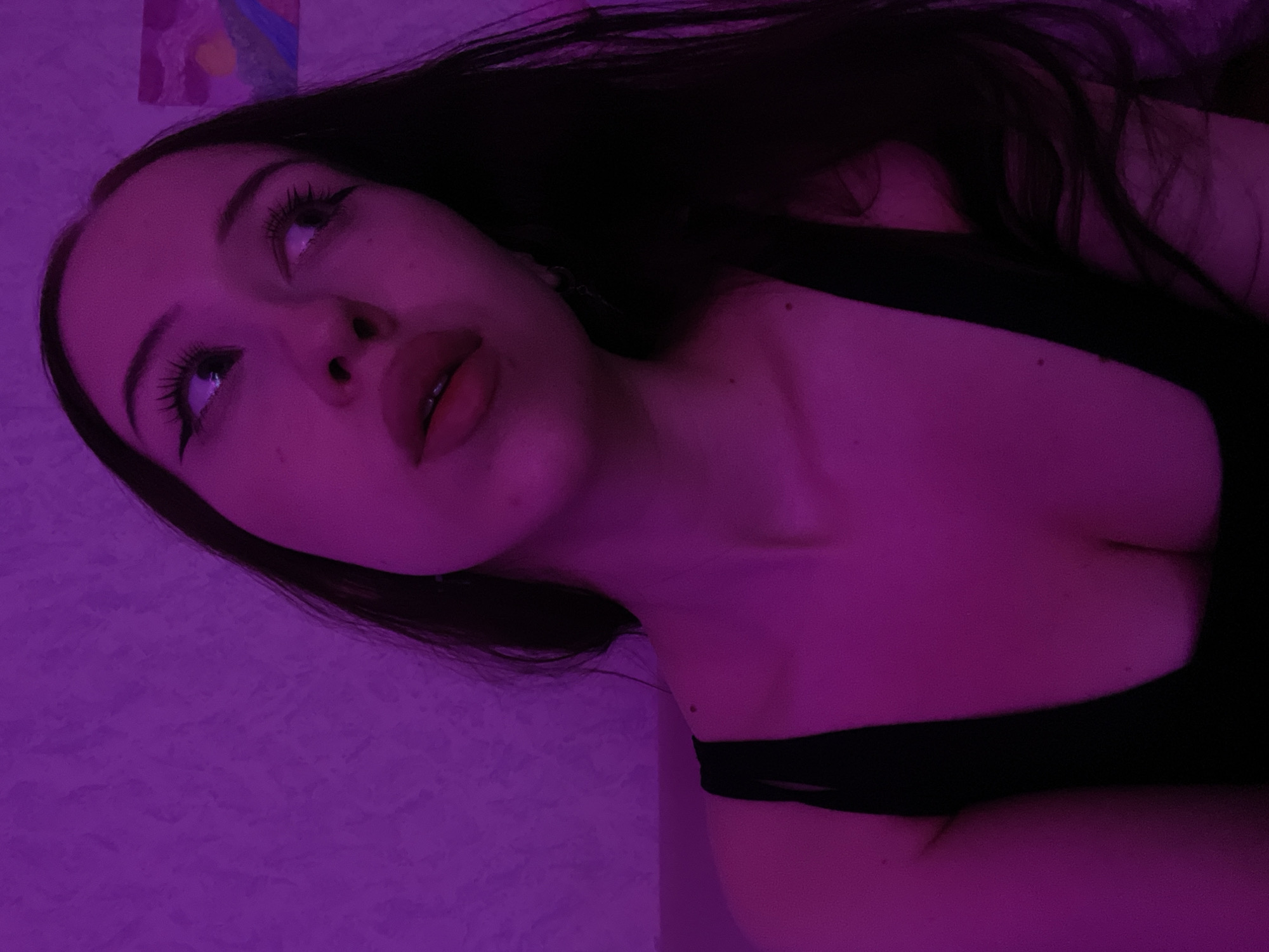 The image shows a close-up of a young woman illuminated by purple lighting. She has dark hair, noticeable eyeliner, and her head is tilted upwards, looking to her right. She is wearing a black top. The background is a textured purple wall with a small abstract painting on the right side. The image does not contain any identifiable landmarks or clues that would suggest a specific location in Votkinsk, Russia.