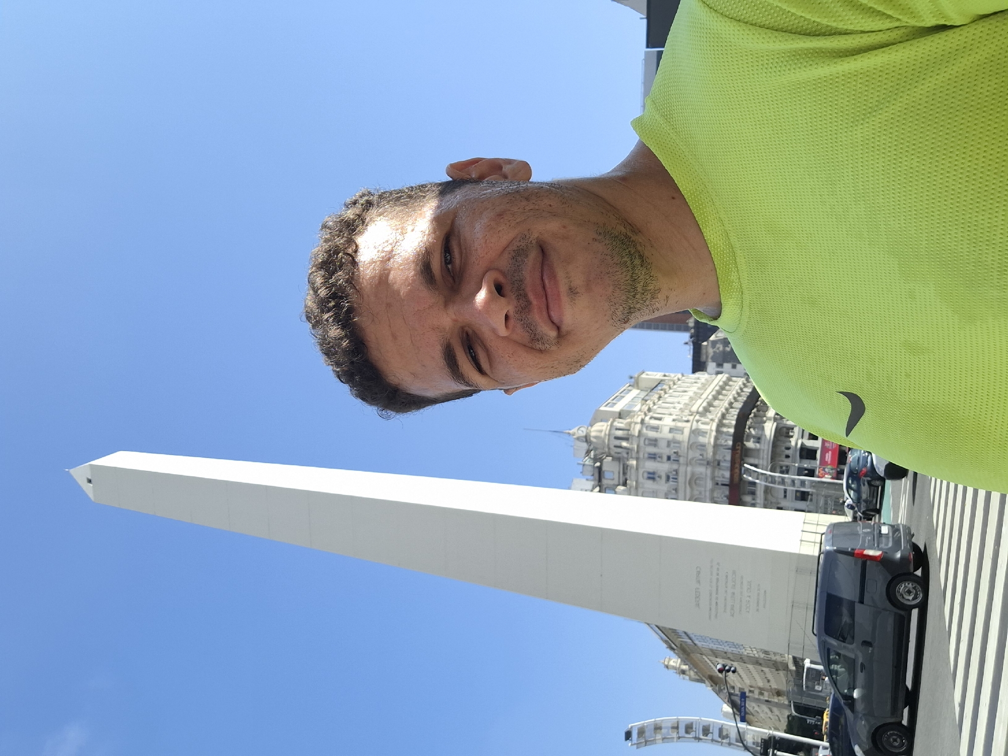 A man is posing for a selfie. The Obelisco de Buenos Aires, a national historic monument in the Plaza de la República in Buenos Aires, Argentina, is visible in the background. A street with vehicles is also visible. The sky is blue. The man is wearing a bright green shirt.