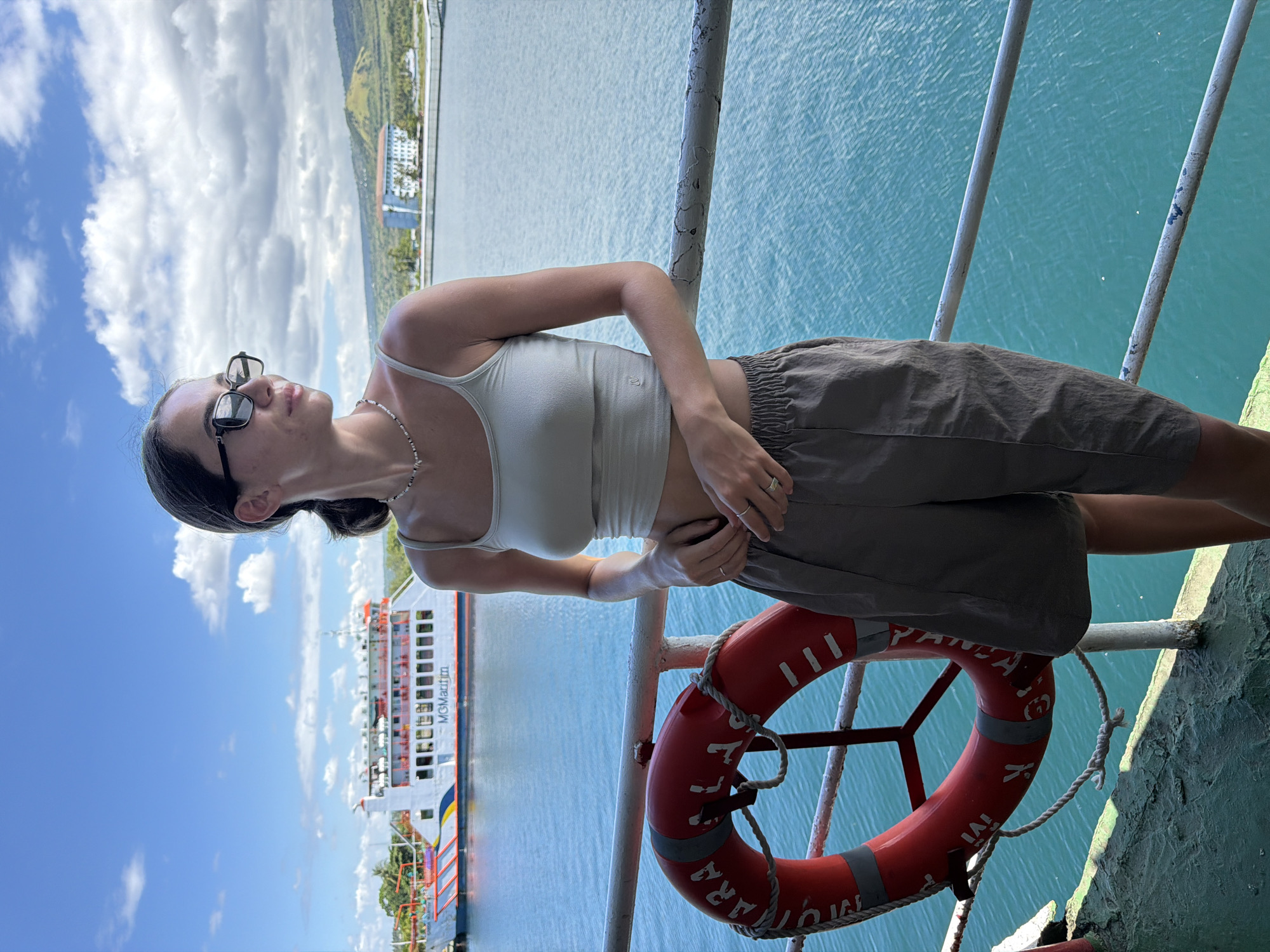 A woman stands on the deck of a ferry, likely in the vicinity of Gunung Malang, Indonesia, given the presence of a vessel labeled "MGMAURITIUS". She is wearing sunglasses, a light-colored tank top, and dark shorts. Her hands are near her stomach. Adjacent to her is a red life preserver. The background reveals a body of water, a shoreline with buildings, and a cloudy sky.