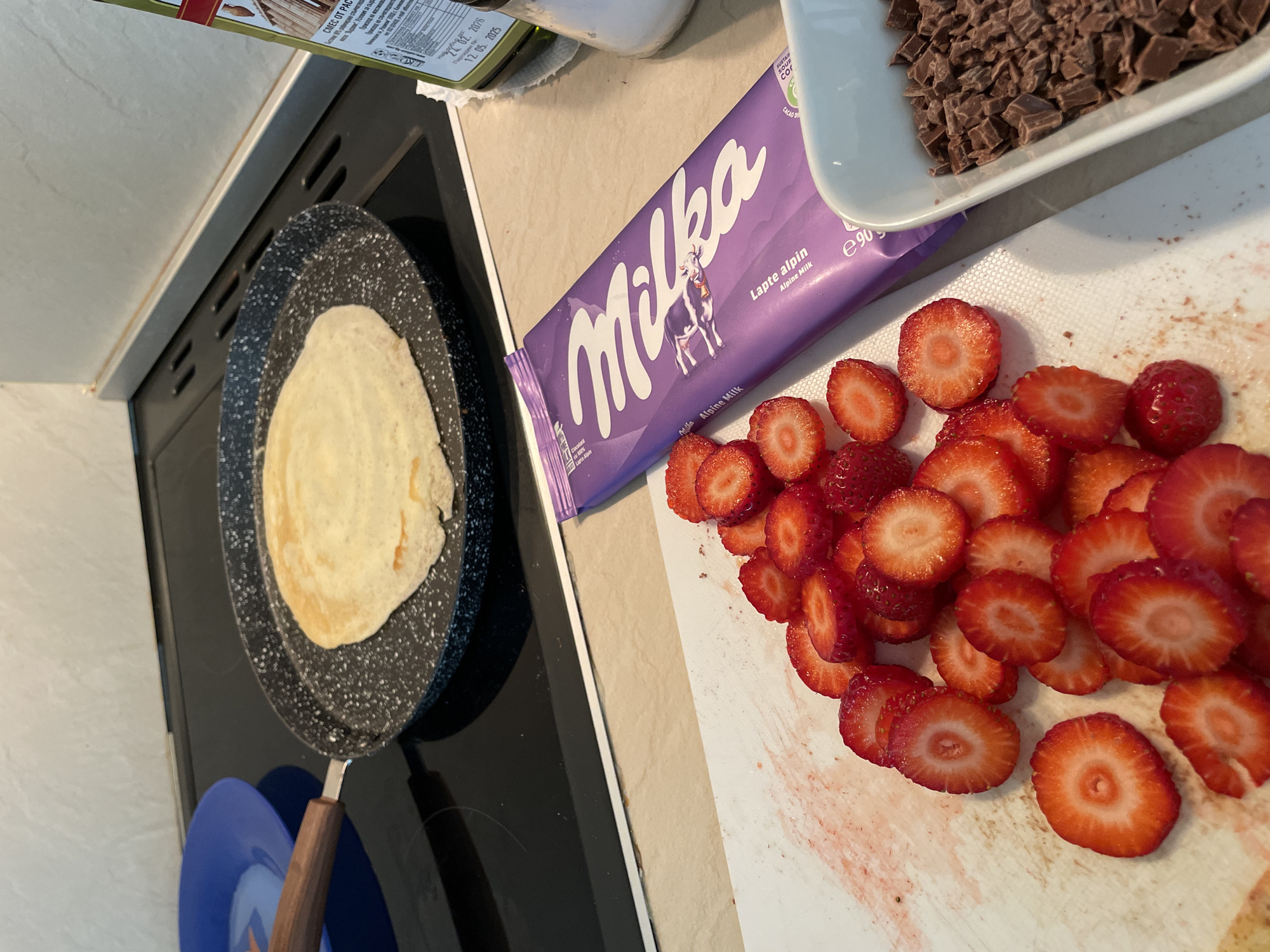 This image captures a close-up, overhead view of a kitchen countertop in Zlatograd, Bulgaria, showcasing ingredients and a pan during food preparation. The central activity involves making crepes or pancakes, with one light-colored pancake cooking in a dark, speckled pan on an electric stovetop in the left foreground.
To the right, a significant portion of the countertop is covered with freshly sliced strawberries, bright red and juicy, scattered across a white cutting board that shows traces of red strawberry juice. Beside the strawberries, a purple Milka "Alpine Milk" chocolate bar lies prominently, its packaging featuring a cow and the brand name. In the upper right corner, a white bowl holds a pile of roughly chopped chocolate pieces, likely derived from a similar chocolate bar, ready for use as a filling or topping.
Partially visible in the top left, an olive oil bottle with Cyrillic text indicates its contents and a visible expiration date of "12. 05. 2025." Other readable text includes "CMEC OT PAC," "кон Верки. Условия за сыр," and "Проксид на маслинов" from the bottle label, and "Milka Lapte alpin Alpine Milk," "Ciocolată Milka Alpine Milk," "e90g," "SUSTAIN," and "CACAO D" from the chocolate bar packaging. A blue plate is subtly visible beneath the handle of the pan. The scene suggests a pleasant domestic activity, likely preparing a sweet meal or dessert.