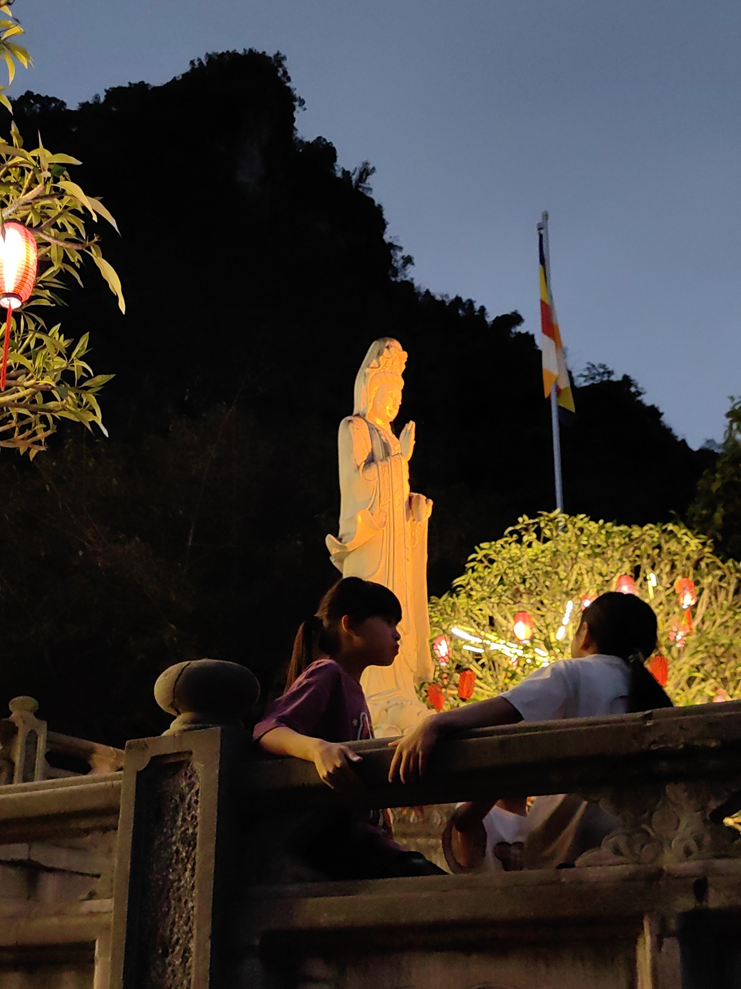 The photo shows a nighttime scene in Xã Đàm Thủy, Vietnam. Two young girls are leaning on a stone railing in the foreground. Behind them is a tall, illuminated statue of a figure with its hands raised in a gesture. In the background, a dark, mountainous landscape rises under a dusk sky. A Buddhist flag is visible to the right of the statue. The scene is decorated with red lanterns. The general atmosphere is one of calmness and possible worship.