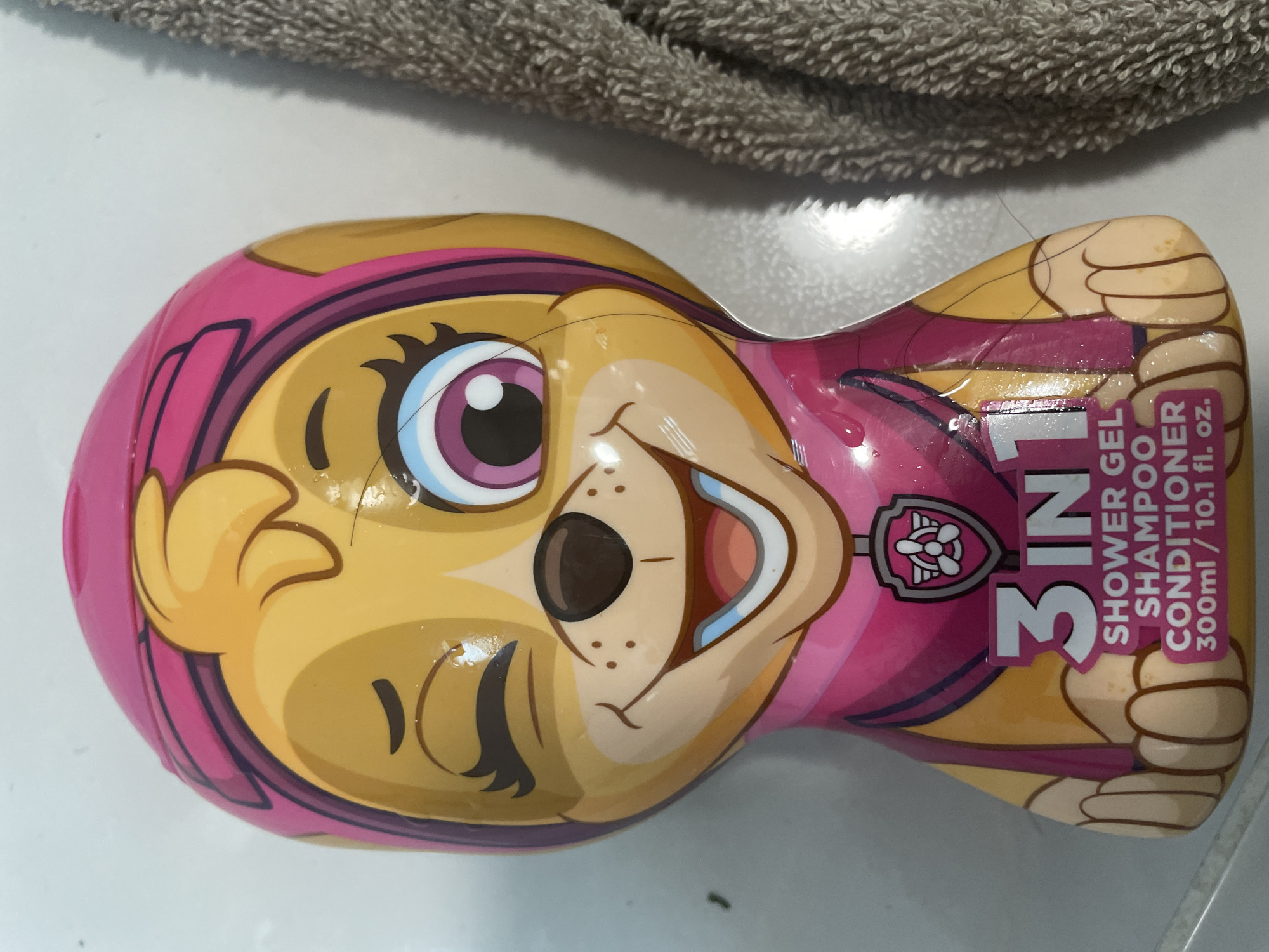 A bottle of "Paw Patrol" themed 3-in-1 shower gel, shampoo, and conditioner is displayed. The bottle features a cartoon character, Skye, a golden retriever puppy wearing a pink aviator hat and goggles. The character is winking and smiling. The product information, "3 IN 1 SHOWER GEL SHAMPOO CONDITIONER 300ml / 10.1 fl. oz." is visible on the lower part of the bottle. A portion of a grey, textured towel is visible to the right of the bottle. The bottle is sitting on a white, tiled surface. The image is likely taken indoors, on a countertop or floor. There is no information in the image to suggest a location in Sevlievo, Bulgaria.