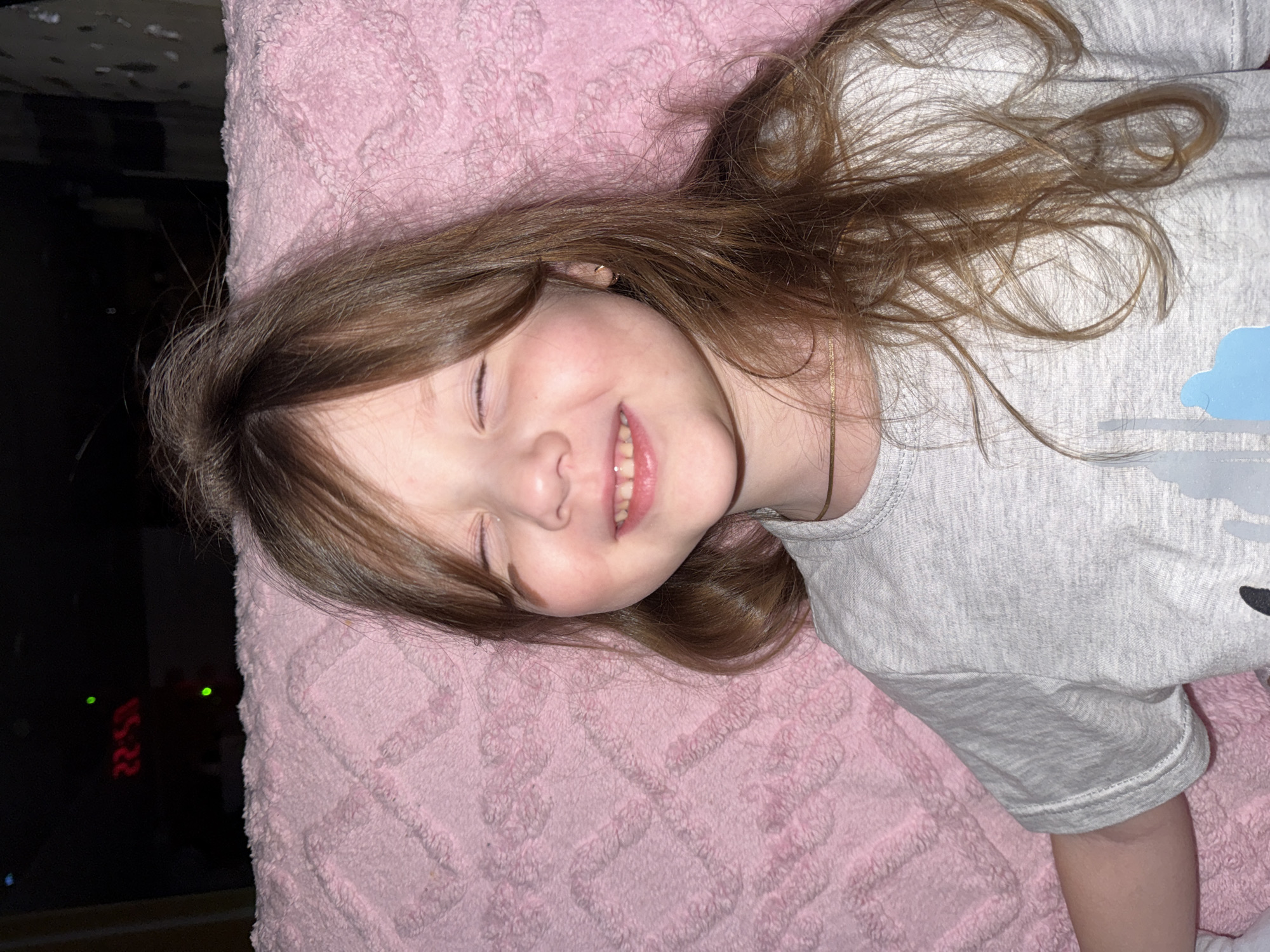 A young girl with long brown hair is lying on a pink blanket, smiling with her eyes closed. She is wearing a gray t-shirt with a blue design on the front. In the background, to the left, there is a dark area with red digital numbers visible, possibly a clock displaying "22:53". The location is not discernible from the image.