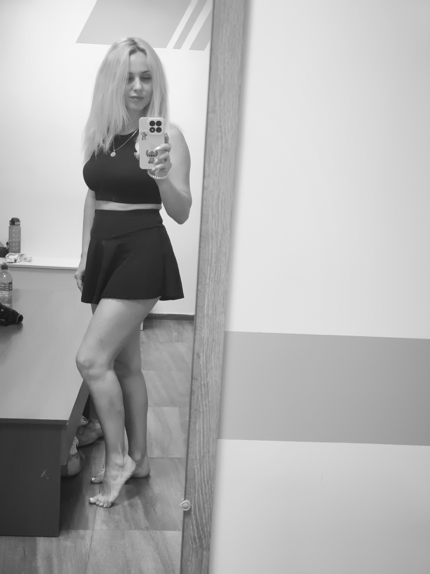 A black and white mirror selfie captures a woman standing indoors, likely in a gym or changing room. She has long, light-colored hair and is wearing a dark sports bra or crop top and a matching dark, flared skirt or skort. She holds a light-colored smartphone with decorative charms, including one resembling Stitch, in her right hand, directing her gaze towards it. She wears a necklace with a round pendant and a beaded bracelet. Barefoot, her right foot is slightly lifted on tiptoes, revealing light-colored toenail polish. To her left, a dark bench or shelf holds a clear water bottle with a dark cap; the label shows a partial, inverted reflection of text, possibly "WEW". The background consists of plain walls, with a horizontal gray stripe on the right, and a wooden or wood-look floor.