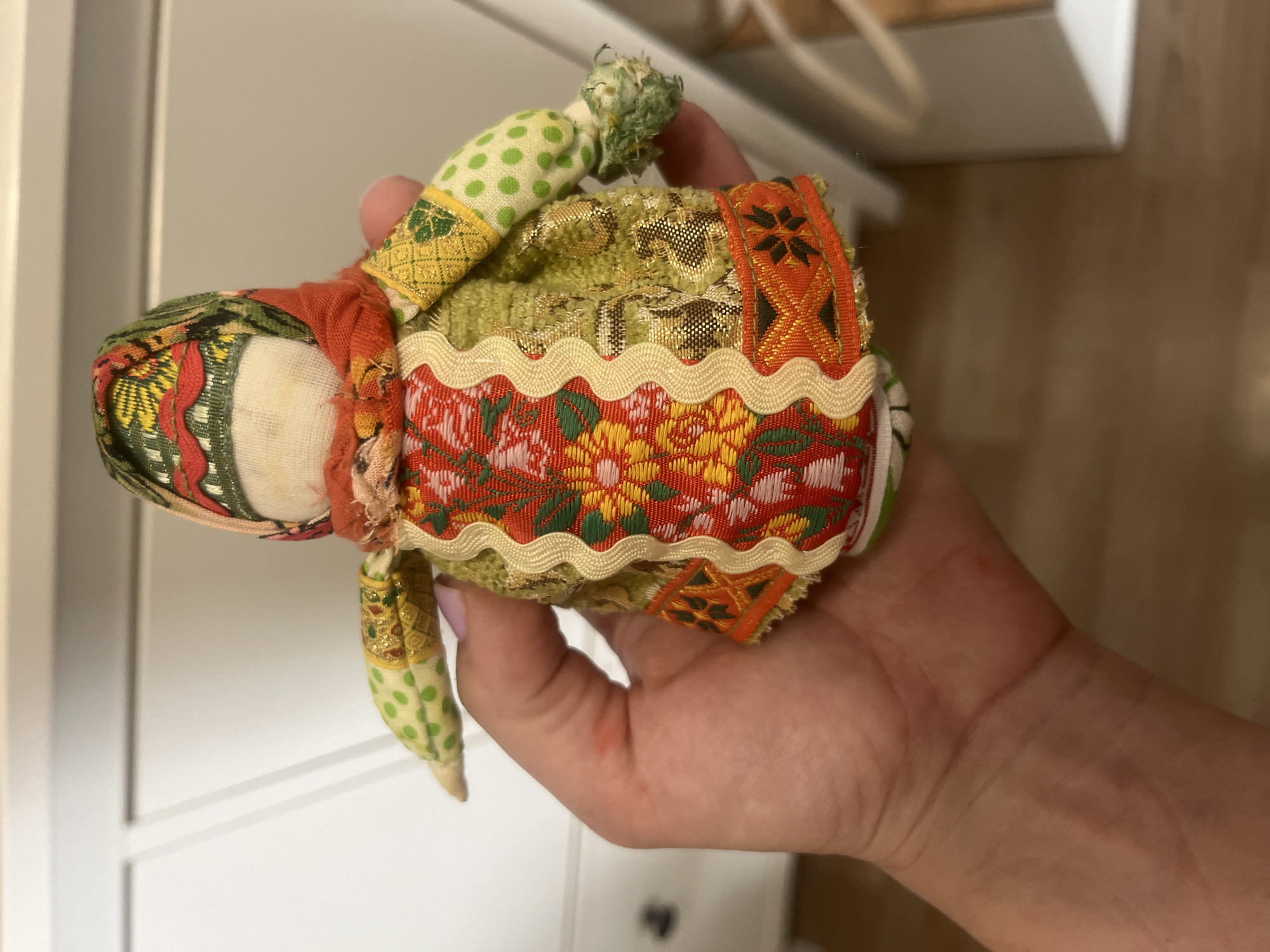 A hand holds a small, intricately patterned fabric doll. The doll is dressed in a vibrant, multi-colored outfit with floral motifs and geometric borders. Its head is covered by a patterned scarf, and it has small fabric arms. The background appears to be an indoor setting, possibly a room in a home, with light-colored furniture. The image does not contain any information or visual cues that could link it to the city of Voronezh, Russia.