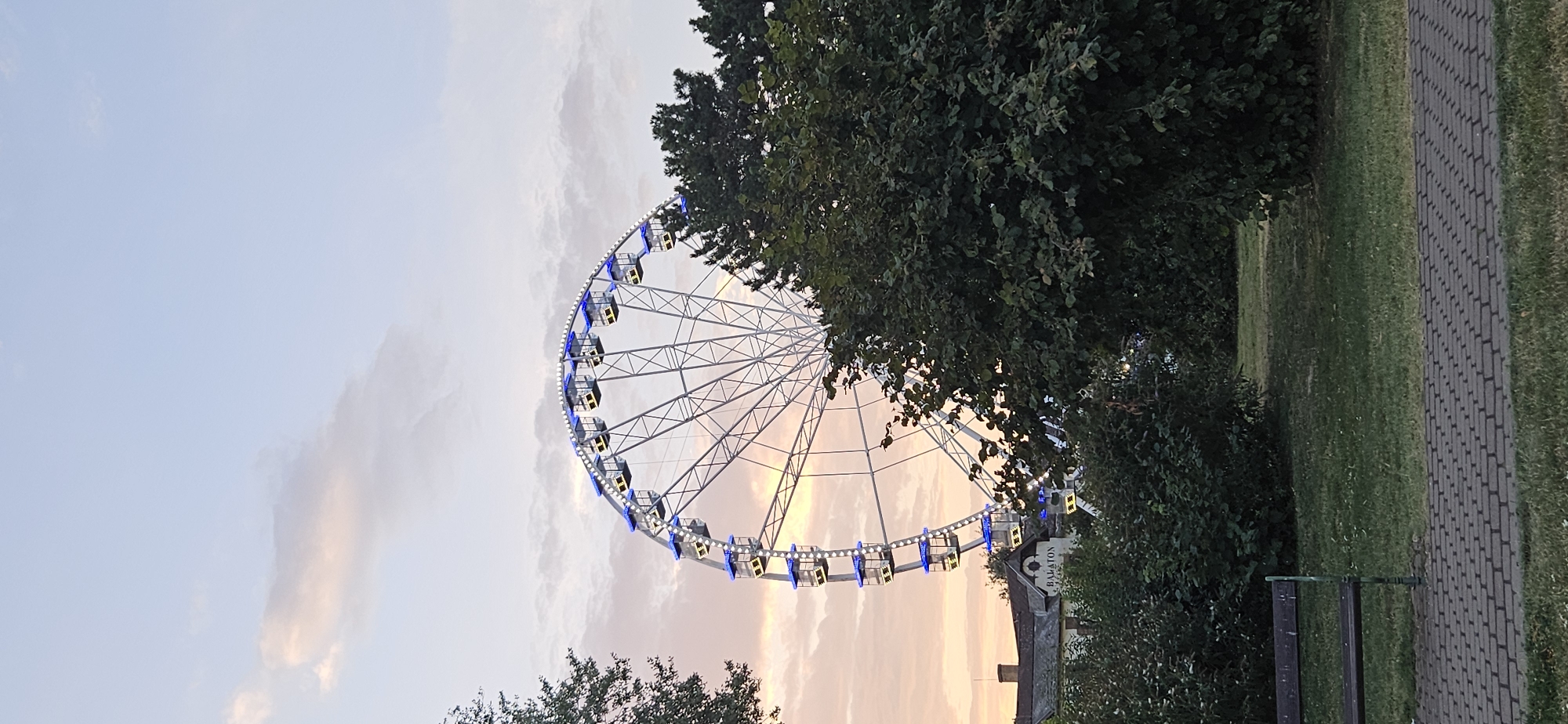 The image shows a Ferris wheel in Keszthely, Hungary, on a cloudy day with the sun setting. The Ferris wheel is illuminated with blue lights and is the main subject of the image. In the foreground, there is a park with a bench and a paved walkway. The Ferris wheel appears to be located near a building with the word "BALATON" visible on it. The overall scene suggests a recreational area or amusement park.