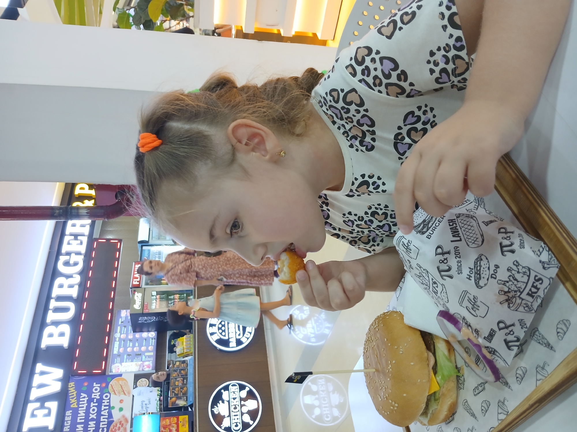 This media file shows a close-up, low-angle shot of a young girl eating in what appears to be a fast-food restaurant. The girl, facing the right side of the frame, is mid-bite into a piece of fried food, possibly a chicken nugget. She has blonde hair tied up with an orange hair tie and wears a white shirt with a black and purple leopard-print heart pattern. Her eyes are focused on the food, and she seems engaged in eating.
In the foreground, on a wooden tray, is a large hamburger with a sesame seed bun, lettuce, cheese, and a slice of red onion. Next to the hamburger is a white paper wrapper with various food illustrations and text. Some visible text includes "BURGER," "LAVASH," "T&P since 2019," "Hot Dog," "KFS," and "Coffee."
In the background, a vertical sign with large white letters reads "EW BURGER." Below it, there are other signs with Russian text, including "АКЦИЯ" (which means "promotion" or "sale") and references to "ПИЦЦУ" (pizza) and "ХОТ-ДОГ" (hot dog). Further in the background, two other people are visible, one standing and one a child wearing a light blue dress. The setting appears to be indoors, likely a food court or a restaurant, with bright lighting. There are no indications of the time of day or weather. The overall mood is casual and focused on the food.