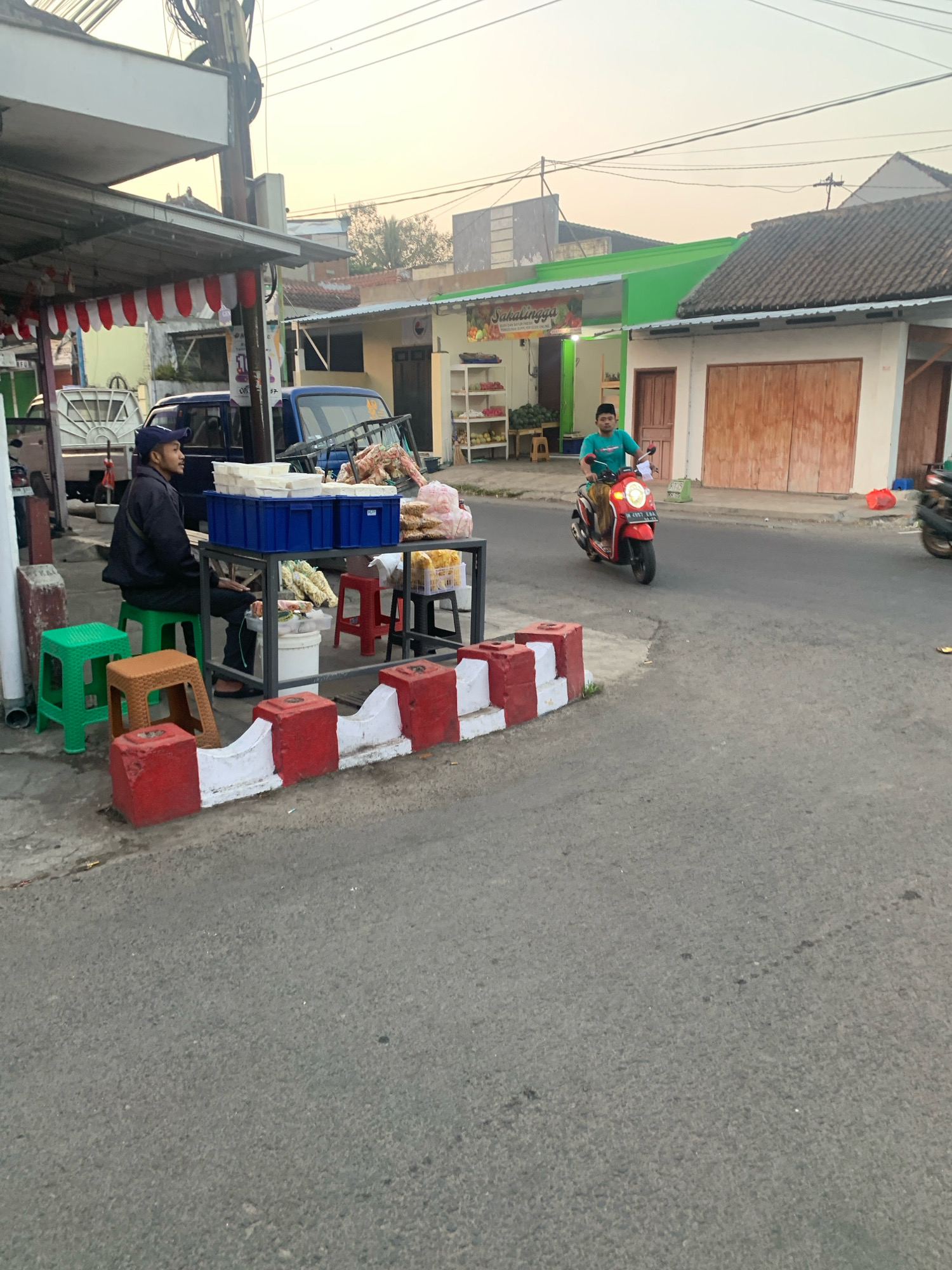 In the city of Bululawang, Indonesia, a street scene unfolds during daylight hours. A man, wearing a dark jacket and a blue cap, sits on a green stool beside a table laden with various food items. He is positioned at a roadside stall, indicated by the red and white barriers lining the pavement. The stall displays stacks of blue containers, bags of packaged snacks, and bundles of what appear to be corn.
Across the street, a young man in a teal shirt rides a red and yellow scooter. He is captured mid-motion, approaching the viewer's perspective. Behind the stall, a blue pickup truck is parked, its bed filled with items. The surrounding environment includes buildings with colorful facades, one painted bright green with a sign that reads "SakaLINGGA," and another with traditional wooden doors. The sky above is a soft, hazy hue, suggesting it is either early morning or late afternoon. The overall impression is one of a typical street scene in a developing urban area, with commerce and daily life intertwining.