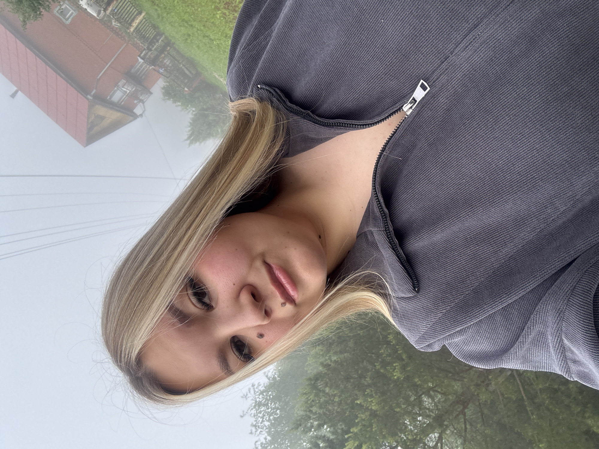 A close-up selfie shows a young person with long, straight blonde hair, wearing a dark grey ribbed zip-up top. Their head is tilted, and they look towards the camera with a neutral expression, revealing moles below one eye and on the upper lip. The zipper of the top is partially open.
The background is an outdoor scene blanketed in mist or fog, suggesting a cool or damp weather condition. On the left side, a reddish-brown building with a prominent red roof is partially visible through the haze. Faint lines, possibly power lines, cut across the misty sky above the building. To the right, green trees are discernible, their forms softened by the fog. The overall setting is rural or suburban, consistent with the location context of Большой Букор, Russia. There is no visible text in the image.