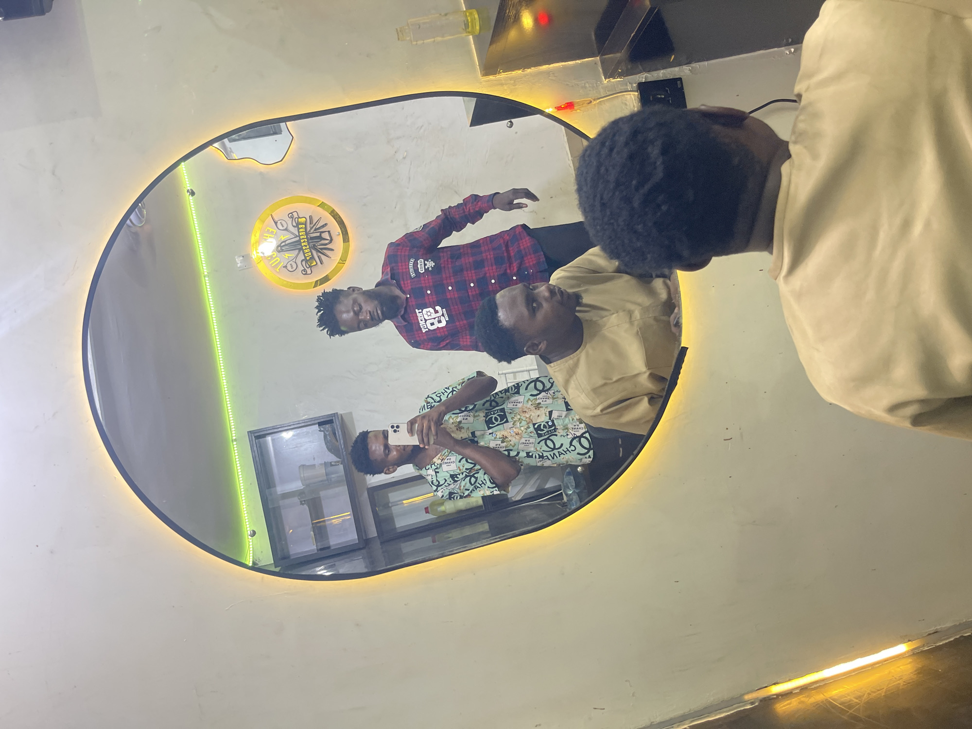 The image captures a mirror, which reflects three men and parts of a barbershop interior. The photo itself is rotated 90 degrees clockwise.
In the foreground, outside the mirror's frame, the back of a person wearing a light brown or tan top is visible, likely the individual taking the photograph.
The prominent feature is a large, irregularly shaped circular mirror, framed by a glowing yellow LED light strip. Within the mirror's reflection, three men are seen. One man, wearing a red and black plaid long-sleeved shirt with a small emblem and a number "85" or "B5" on the sleeve, stands looking downwards. Beside him, a second man in a light brown or tan short-sleeved top is seated, looking towards the man in plaid. A third man, seated to the left of the others in the reflection, wears a light green patterned shirt that appears to feature "CHANEL" logos. He is holding a smartphone up, seemingly taking a picture of himself and the others in the mirror.
The reflected background behind the men includes a light-colored wall, a vertical green LED light strip, and a circular yellow sign. The text on the sign, reflected backward, reads "THE BARBER SHOP" or a similar phrase, accompanied by illustrations of barber tools like scissors and combs. A small glass display case and a bottle are also reflected. The setting appears to be a modern barbershop, indicated by the mirror design and accessories, illuminated by various artificial lights. The scene suggests a casual social moment in Unguwar Gabas, Nigeria.