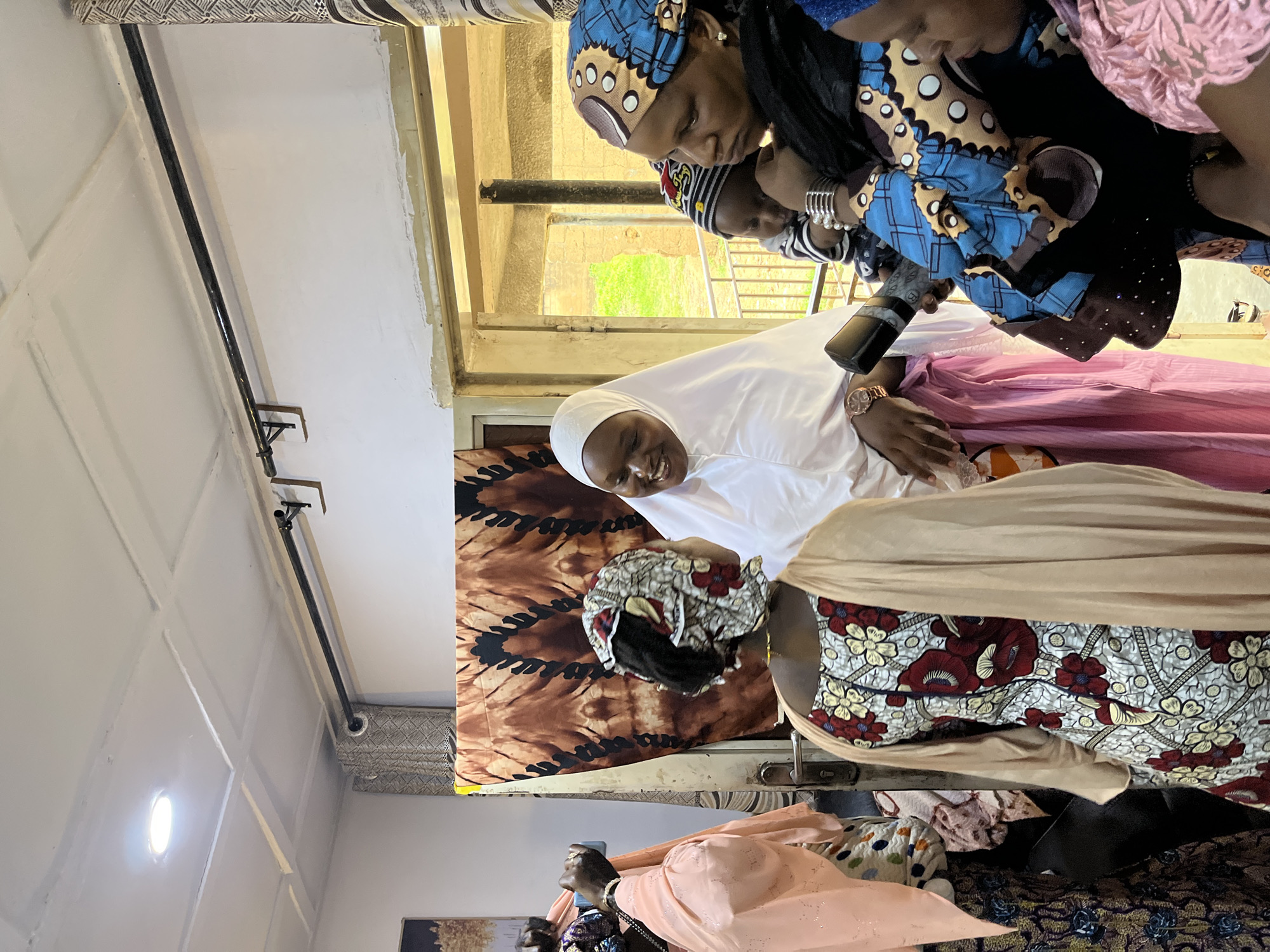 The image depicts a gathering of individuals inside a room, likely in Barakin Akawo, Nigeria. The central focus is on a group of women, some of whom are wearing traditional African attire with vibrant patterns and headscarves. One woman, in the center, is wearing a white hijab and a pink skirt, and appears to be smiling and engaged in conversation. To her right, another woman in a blue and gold patterned headscarf holds a baby. Another woman, wearing a light pink outfit and a headscarf, is holding a cell phone, possibly taking a picture. Behind them, a large piece of patterned fabric hangs as a backdrop. The room has a white ceiling with recessed lighting and a dark rod mounted on the wall, likely for curtains. The overall scene suggests a social or familial gathering.