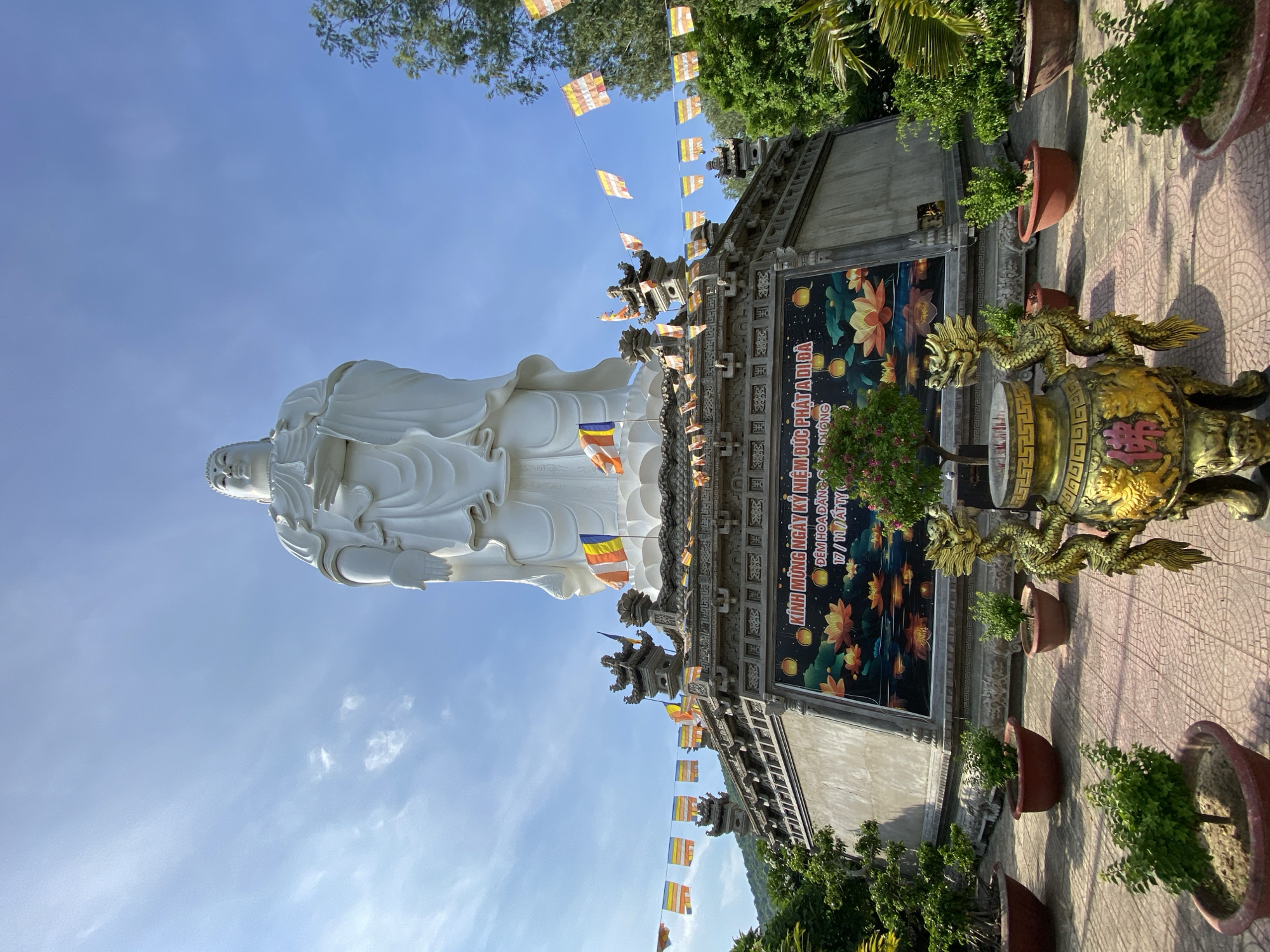 The image depicts a large, white statue of a Buddha, likely Quan Am Bodhisattva, set against a bright blue sky with scattered clouds. The statue stands atop a multi-tiered base, adorned with decorative stonework and numerous colorful prayer flags strung across its upper levels. A large banner with floral imagery, specifically lotus flowers, and Vietnamese text is displayed on the front of the base. In the foreground, a large, ornate golden incense burner with a dragon motif is positioned on a paved courtyard. Several potted plants and trees are arranged around the courtyard, with lush green foliage contrasting with the red-tiled ground. The overall scene suggests a spiritual or religious site, possibly a temple or monument, located in a warm, outdoor environment. The presence of Vietnamese text on the banner and the style of the religious iconography suggest a location in Vietnam. Specifically, this statue is a prominent landmark in the Tay Nha Trang Ward of Tay Ninh City, Vietnam.