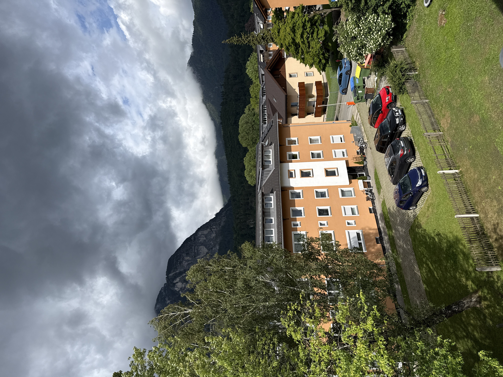The image presents a high-angle view of a residential area in Garmisch-Partenkirchen, Germany, with a dramatic mountainous backdrop under a heavily clouded sky. The upper two-thirds of the scene are dominated by dense, dark grey cumulonimbus clouds, though small patches of brighter sky are visible to the right. Majestic dark grey mountains, characteristic of the Bavarian Alps, rise in the background, their peaks partially shrouded in mist and low-hanging clouds. The lower slopes of the mountains are covered in dark green coniferous forests.
In the midground, several multi-story residential buildings are visible. The most prominent structure in the left-center is an orange-colored building with numerous white-framed windows, some featuring white external rolling shutters. It has a dark sloped roof with multiple dormer windows. Other buildings in the midground and right feature brown sloped roofs, many equipped with solar panels, and facades ranging from light yellow to beige, some with wooden balconies.
A paved parking area in the foreground and midground displays several vehicles: a dark blue compact car (left), a black SUV and a red SUV parked side-by-side (center-right), and a light blue sedan positioned behind a white barrier arm (further back). Two green and one yellow waste bin are situated near the light blue sedan. Two bicycles are leaning against the wall of the orange building on the right.
The scene is rich in vegetation, including a large deciduous tree with vibrant green foliage on the left, partially obscuring the orange building. Other smaller trees, bushes, and a tall, slender conifer are dispersed throughout the residential spaces. A patch of green grass with a rustic wooden picket fence crosses the bottom of the image. No people, specific actions, or interactions are visible.