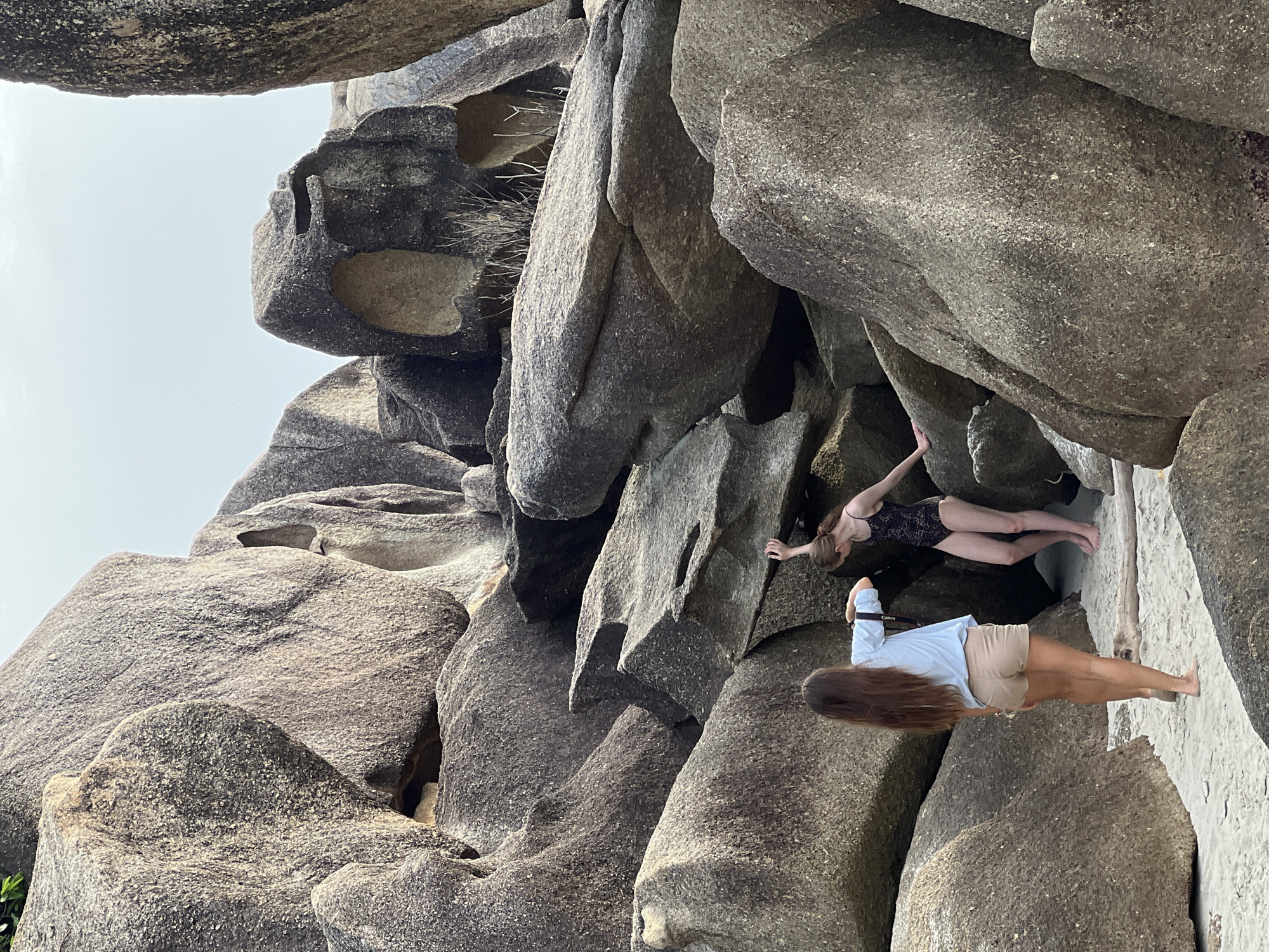 Two individuals are on a sandy beach amidst large rock formations. The person on the left, viewed from the back, has long brown hair and is wearing a light blue t-shirt and tan shorts. They are holding a phone up, seemingly taking a picture. The person on the right is wearing a black one-piece swimsuit and is facing away from the camera, looking towards the rock formations. They have their right arm extended upwards, touching a rock. The environment appears to be a rocky beach with a pale sky overhead, suggesting the location is Baan Chaweng Noi, Thailand.