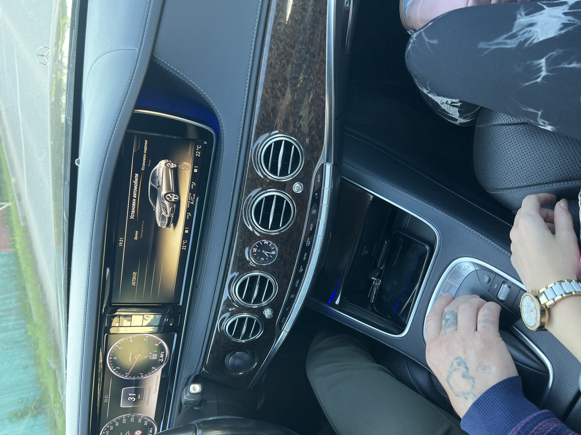 The image shows the interior of a Mercedes-Benz S-Class, likely in motion, as indicated by the speedometer reading of 31. The car is equipped with a large central infotainment screen displaying the car's settings and a digital clock showing 19:31. The driver's hand, with visible tattoos, is resting on the center console's rotary dial, suggesting they are operating the infotainment system. A passenger's hand, wearing a watch, is also visible. The dashboard features wood trim and climate control vents. The scene is consistent with a vehicle interior in an urban or suburban environment, with the bright daylight and greenery visible through the windshield suggesting it's daytime. The context of the image being taken in Krasnoe-na-Volge, Russia, provides a geographical setting for this domestic scene.