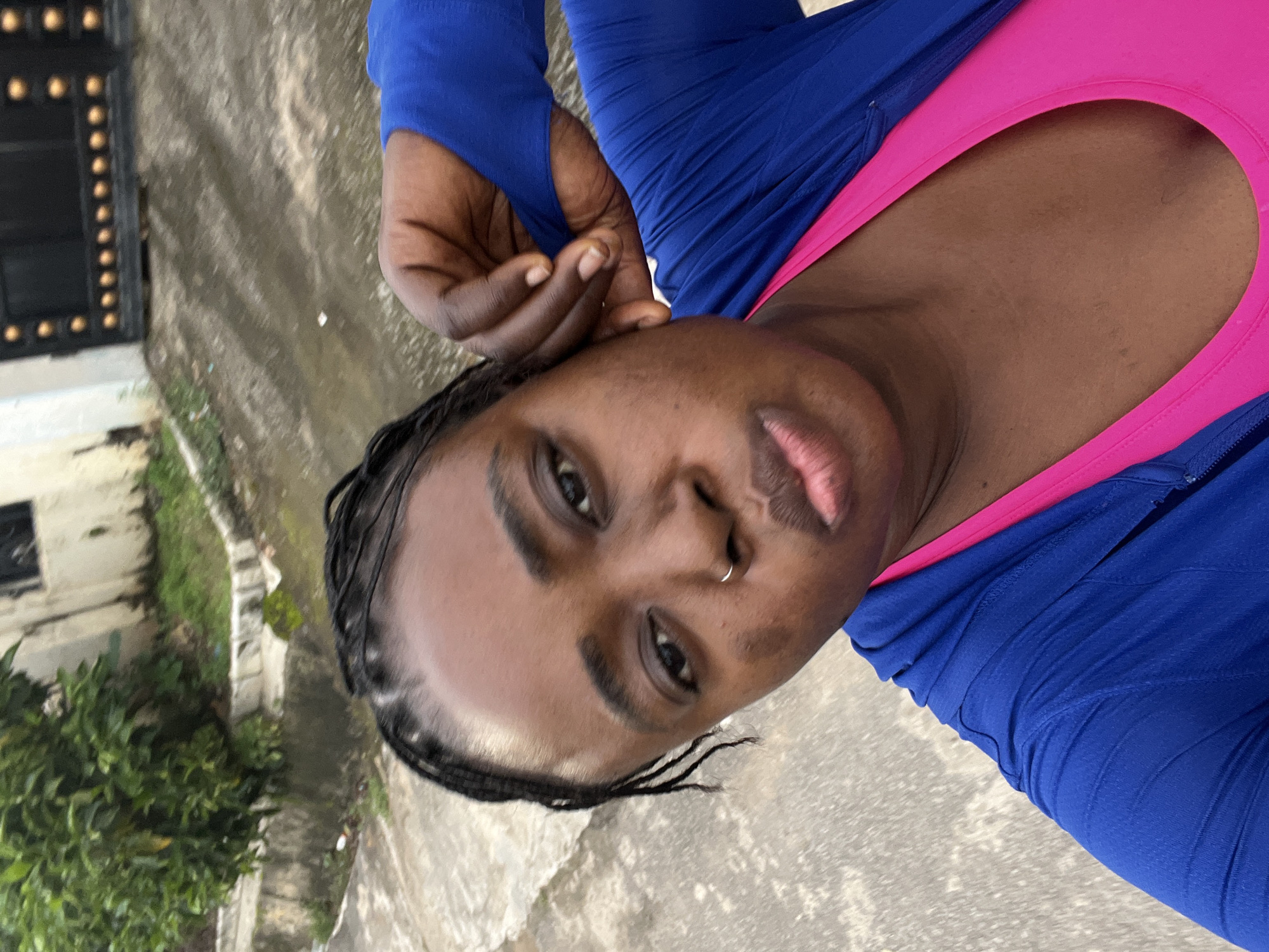 The image is a close-up, waist-up selfie of a Black woman outdoors in Abuja, Nigeria. She is wearing a bright pink sports bra and a blue zip-up jacket with the hood up. Her dark hair is braided and pulled back. She has a nose ring and is posing with her right hand near her ear, looking at the camera with a slight pout. The background shows a paved area with some greenery and a building with a decorative metal gate. The lighting suggests it is daytime.