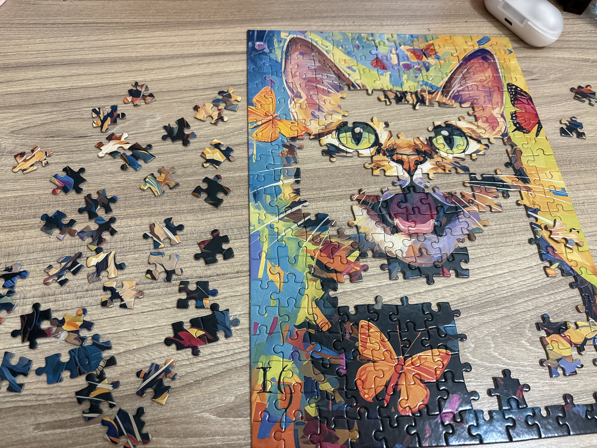 The image shows a partially completed jigsaw puzzle of a cat with butterfly motifs. The puzzle is on a light-brown, wood-grain surface. The cat's face is largely complete, but sections like the top of the head and the areas around the eyes are missing pieces. A collection of loose puzzle pieces sits to the left. A white earbud case is in the upper right corner.