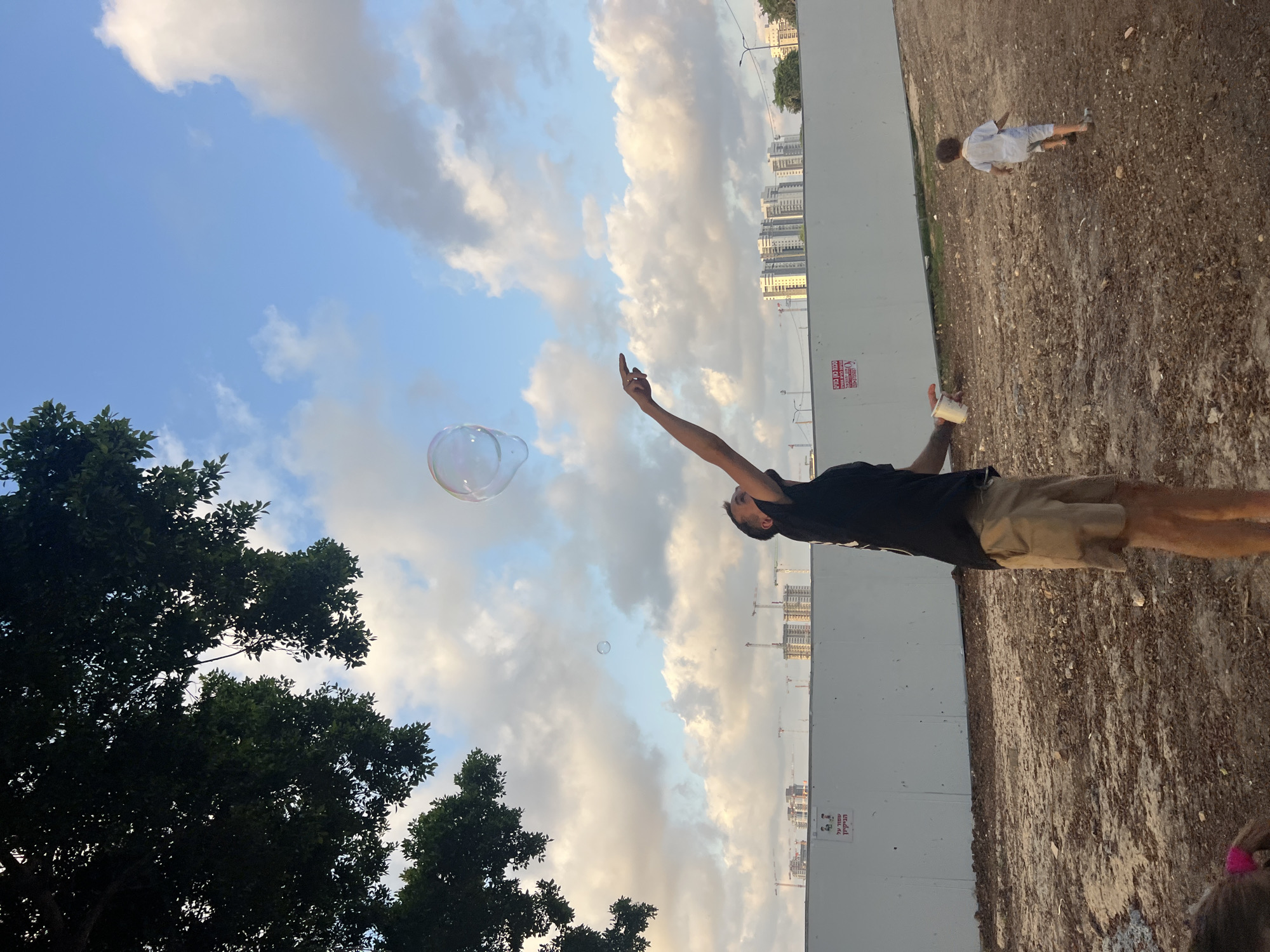 A man in a black t-shirt and khaki shorts is blowing bubbles outdoors. He is captured mid-action, with one arm raised and his index finger pointing towards a large bubble floating in the sky. Another, smaller bubble is also visible further away. The sky is a mix of blue and cloudy, suggesting it might be late afternoon or early evening.
In the background, a child in a white shirt and blue shorts is walking on a dirt path. A white construction barrier stretches across the middle ground, behind which a cityscape with tall buildings and construction cranes is visible. The ground is covered in dirt and debris.
A large, dark green tree dominates the left side of the frame, its branches reaching towards the sky. The overall scene appears to be a casual outdoor activity, possibly in a park or an undeveloped area near a city.
Visible text on the white barrier reads "שטח פינוי" which translates to "Evacuation area" and "נא לא להיכנס" which means "Please do not enter." The location is identified as Kiryat Bialik, Israel.