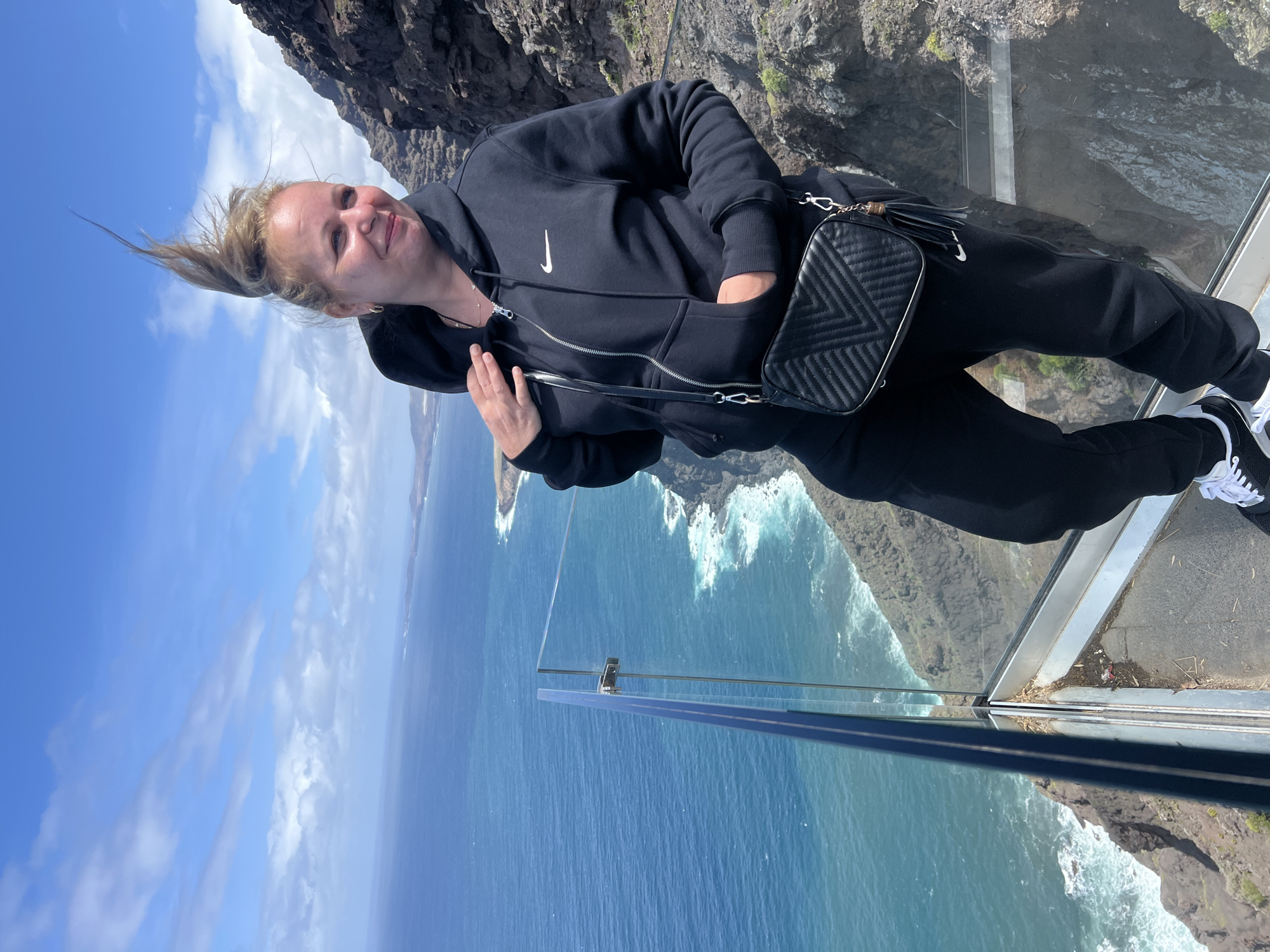 A woman in a black hoodie, sweatpants, and black and white sneakers stands on a glass platform, likely a viewpoint, at Roque del Agua Dulce, Tirma, Spain. She has a black handbag with a crossbody strap. The background includes the ocean, cliffs, and blue sky with scattered clouds.