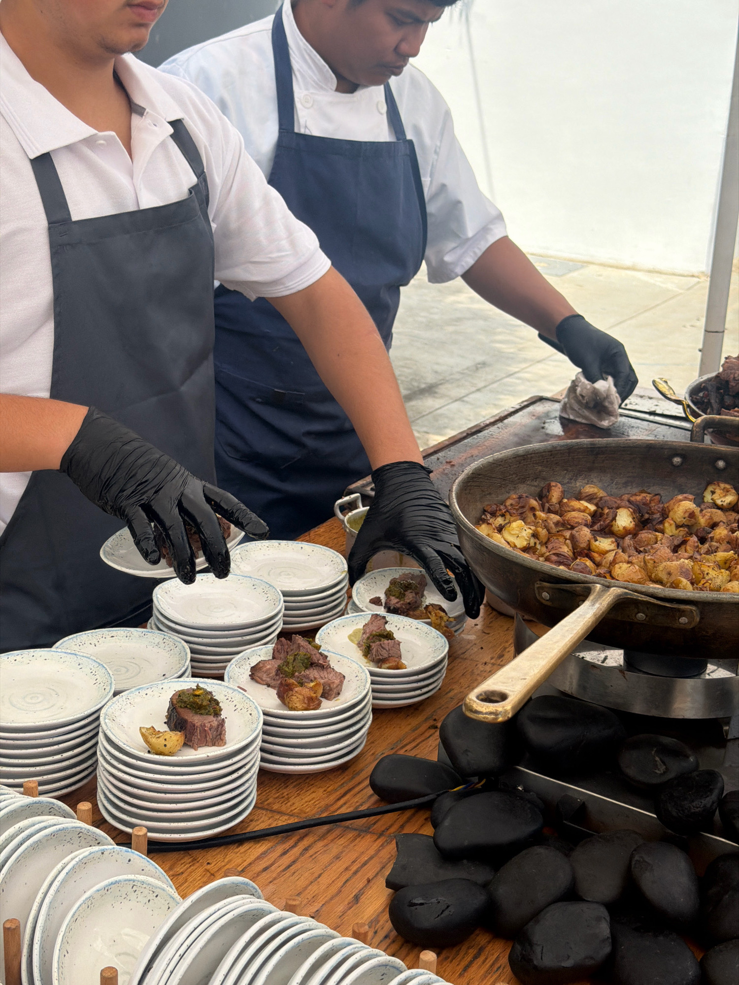 In Kadima, Israel, two chefs in white chef coats and black aprons are preparing food outdoors. The chef on the left, wearing a black apron over a white collared shirt, is using black gloves to place a piece of meat and a potato onto a stack of small plates. The chef on the right, also wearing black gloves and a dark blue apron, is wiping their hands with a cloth. In front of them, a large cast-iron skillet filled with roasted potatoes sits on a stovetop. Several stacks of plates, some with food already on them, are arranged on a wooden table. The background is blurred, but appears to be an outdoor setting with a light-colored wall.
