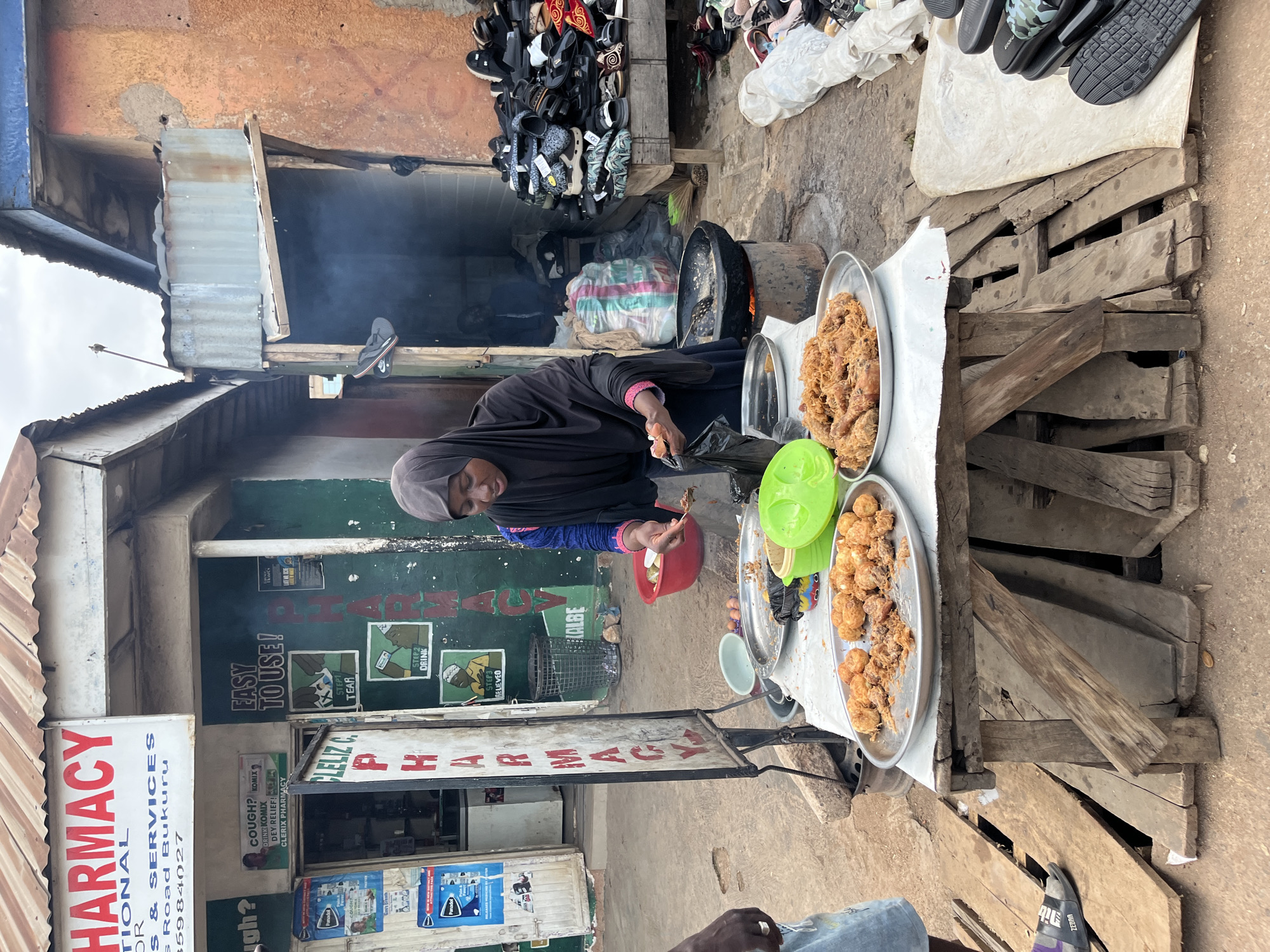 A street scene in Bukuru, Nigeria, features a woman in a black hijab and blue and pink patterned tunic standing behind a table laden with fried food. She is in the process of transferring food from a frying pan to a plate, holding a spoon in her right hand and a plastic bag in her left. In front of her, on the table, are several trays filled with golden-brown fried items, some appearing to be chicken pieces. A red bowl filled with a light-colored food item and a ladle sits next to the frying station. The scene is set against a backdrop of market stalls. To the left, a pharmacy sign reads "PHARMACY" in large red letters, with smaller text below indicating "Drs & Services, Pius Road, Bukuru." Another sign for "CL ELIZ C. PHARMACY" is also visible. To the right, a stall displays a large collection of footwear, including sandals and flip-flops. Smoke rises from the frying pan, suggesting active cooking. The ground is rough and unpaved, typical of an outdoor market.