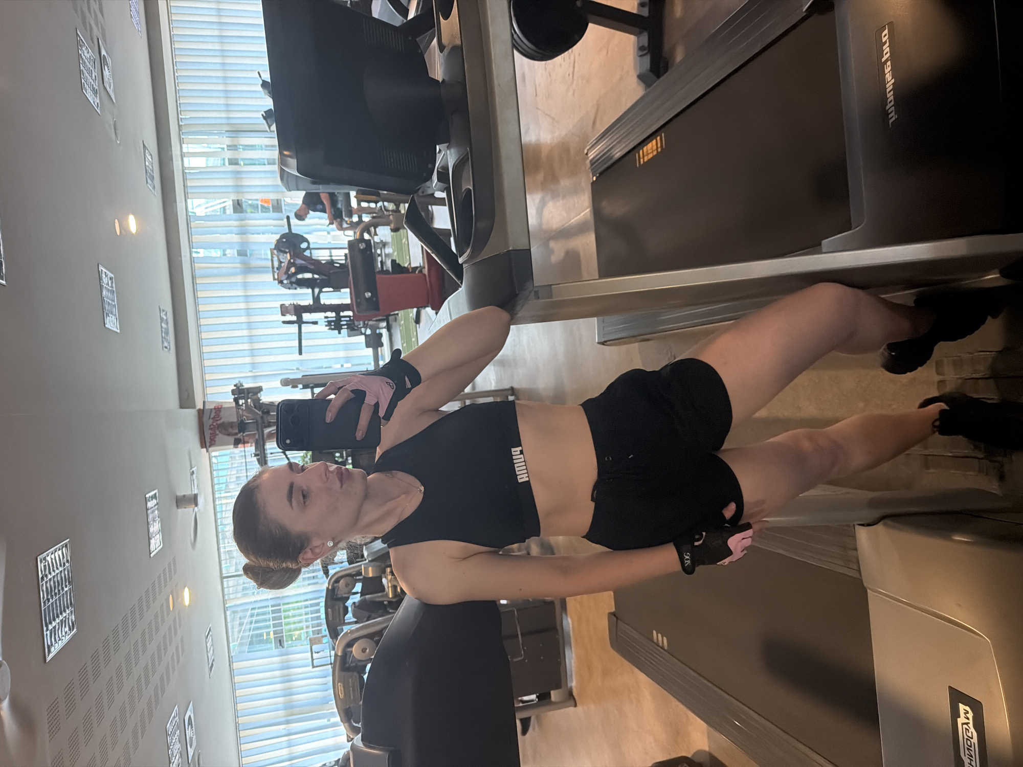A young woman with her hair pulled back in a bun is taking a mirror selfie in a gym. She is wearing a black sports bra and black athletic shorts, with pink and black weightlifting gloves on her hands. She is standing next to a treadmill, and other gym equipment, including treadmills, weight machines, and a person sitting on a weight machine, are visible in the background. The gym appears to be well-lit, with fluorescent lights on the ceiling and natural light coming from a large window. The overall impression is of a modern fitness facility.