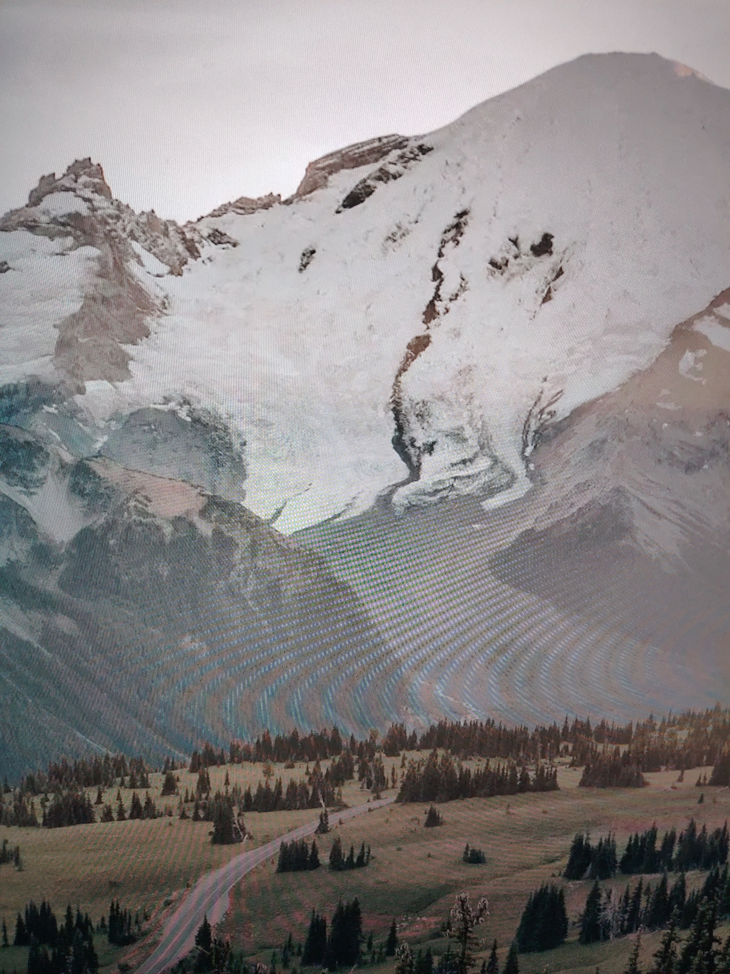 The image shows a mountain range with a snow-covered peak. A valley with evergreen trees is at the base of the mountain. A road cuts through the valley. The location is unknown.