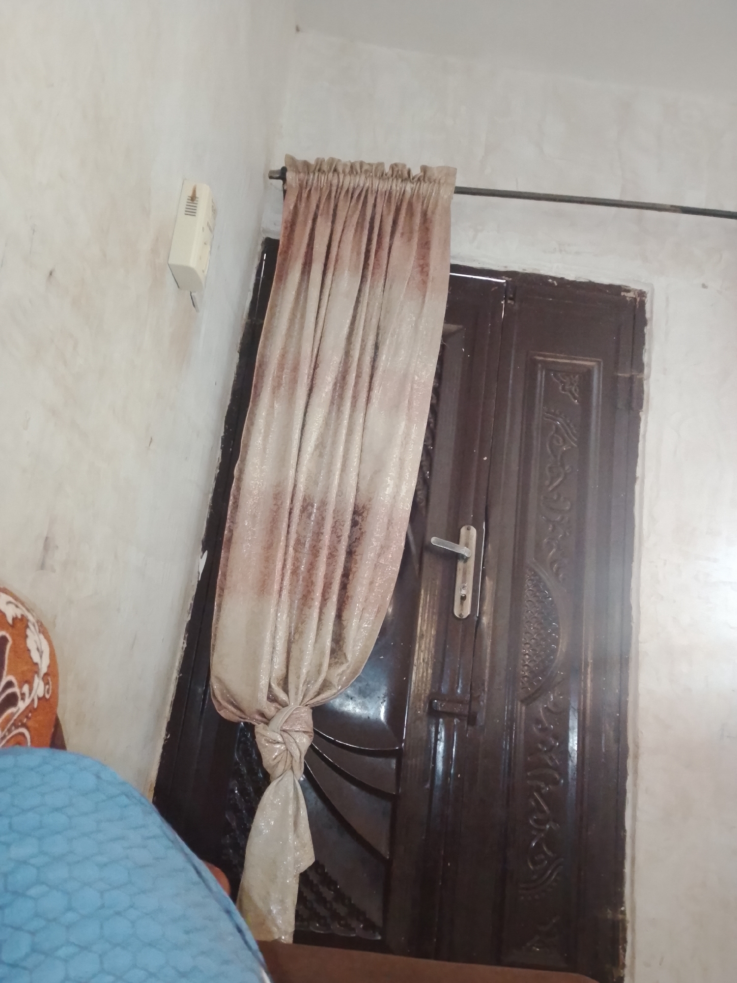 The image shows a close-up view of a dark wooden door in what appears to be an indoor setting. The door has an ornate design carved into its surface, with repeating patterns. A silver door handle is visible on the right side of the door. To the left of the door, a draped curtain with a shimmering, textured fabric in shades of beige and pink is tied back with a knot. The curtain is hung from a dark rod.
On the wall to the left of the curtain, a small, rectangular white object is mounted. It has vertical slats and appears to be some kind of electrical fixture or outlet. The wall itself is a light, off-white color and has a slightly textured finish, with some faint darker marks or smudges. In the bottom left corner of the image, a portion of a blue object with a hexagonal pattern is visible, possibly furniture or upholstery. Another part of an orange object with a patterned design is also partially visible in the lower left.
The lighting in the image suggests an indoor environment, possibly during the day, but there are no strong indicators of time of day or weather. The scene is static, with no apparent activity taking place. The overall impression is of a simple, functional room with some decorative elements. There is no visible text in the image. The location context is Jalingo, Nigeria, but this information is not directly evident from the visual cues in the image itself.