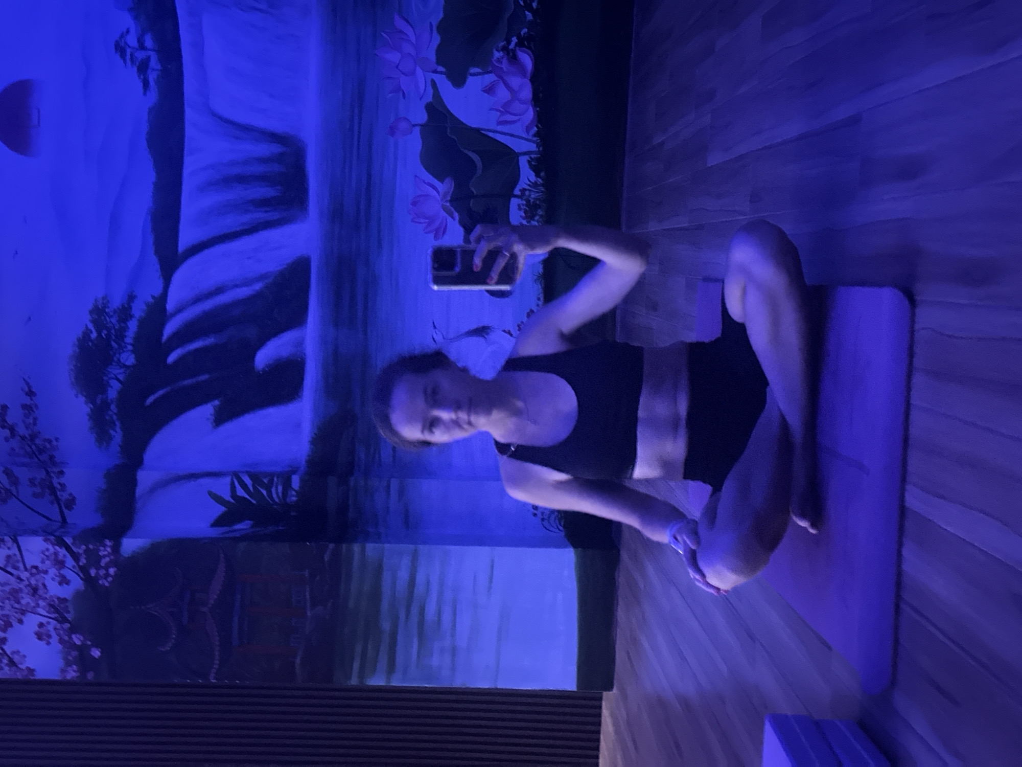 A woman is seated in a cross-legged position on a purple yoga mat in a room with a mural. She is wearing a black sports bra and black shorts, and is holding a phone up, presumably taking a selfie. The background mural depicts a waterfall, lotus flowers, trees, and a temple, illuminated with blue light. The flooring is wooden. The location is identified as Bac Nha Trang Ward, Vietnam.