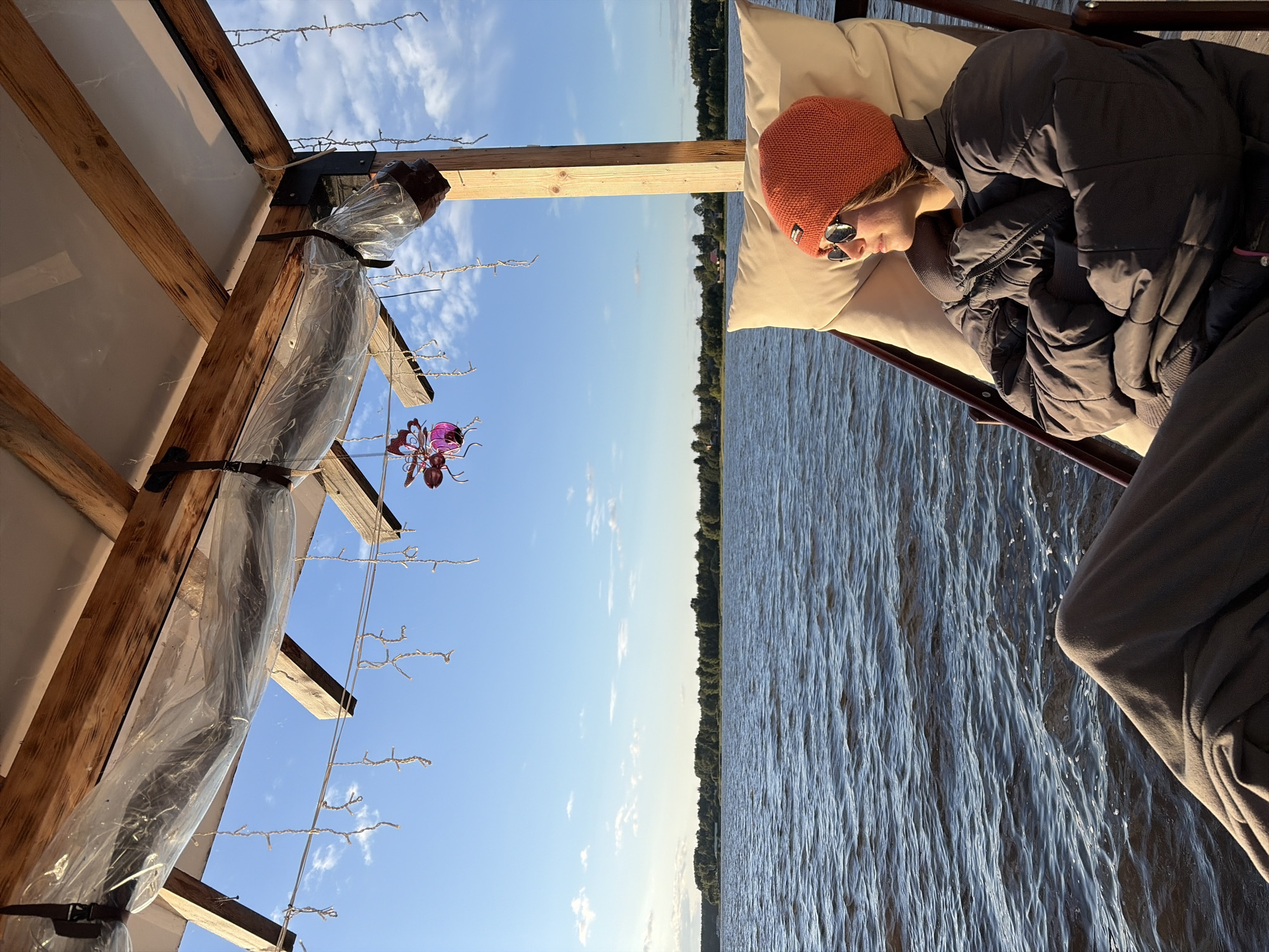 A person sits in a lounge chair on a wooden structure overlooking a body of water. The person is wearing an orange beanie, sunglasses, a grey puffer jacket, and dark pants covered by a grey blanket. The person is facing towards the right of the frame, looking out at the water. The water is choppy, with ripples and small waves. The background shows a tree line with some houses, under a blue sky with scattered clouds. Hanging from the wooden structure are strings of lights and a pink decorative object. The location is the city of Vidogoshchi, Russia.