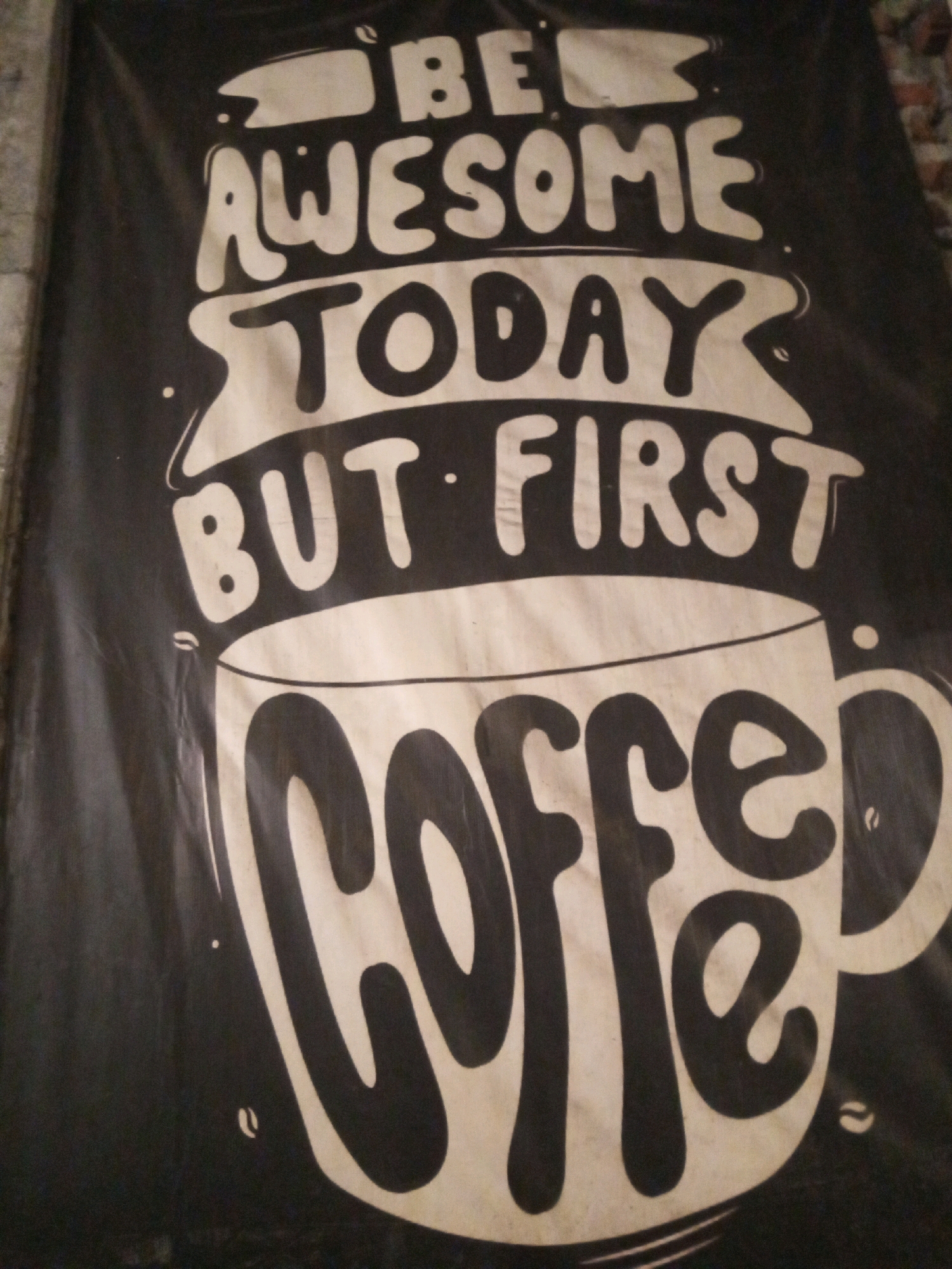 This is a close-up shot of a black fabric banner with white lettering. The banner displays a motivational quote about coffee, with large, stylized text. The top line reads "BE," followed by "AWESOME TODAY," and then "BUT FIRST." The bottom portion of the banner features a large drawing of a coffee cup with the word "COFFEE" written inside it in a thick, rounded font. The image is slightly tilted. The banner is displayed outdoors, with a textured, brick-like wall visible in the background on the left side. The lighting suggests it is daytime, and the overall scene appears to be in a public or commercial area, possibly a cafe or street stall. Given the context of Tambakagung, Indonesia, this banner likely advertises or promotes coffee in a local establishment.