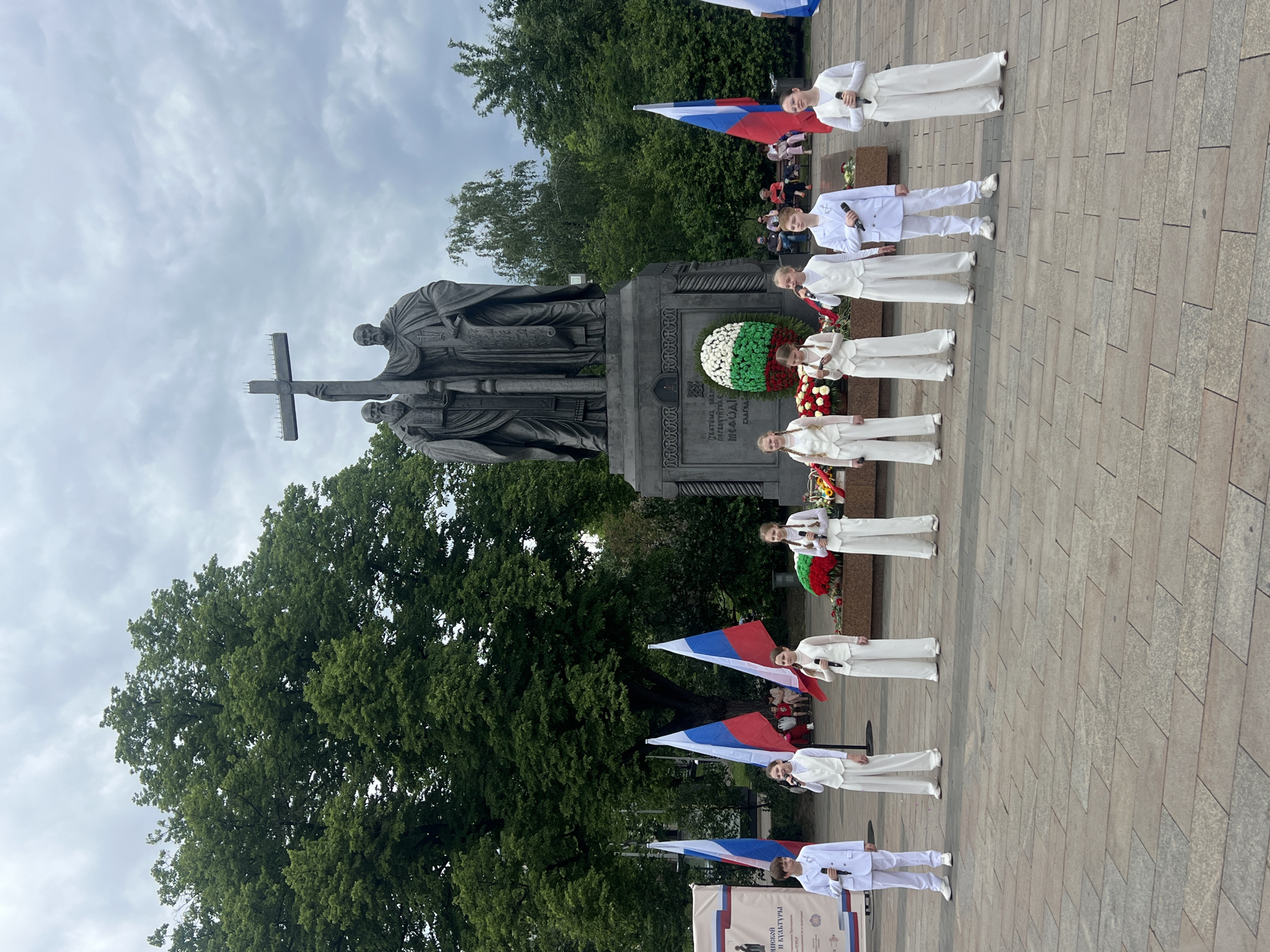 The image shows a statue in Moscow, Russia, likely a monument to saints, with a cross in the foreground. A group of young people in white clothing is lined up in front of the statue. They each have a microphone and some are holding Russian flags. They appear to be singing or performing a ceremony. In the background there are trees and an overcast sky. The area is paved.