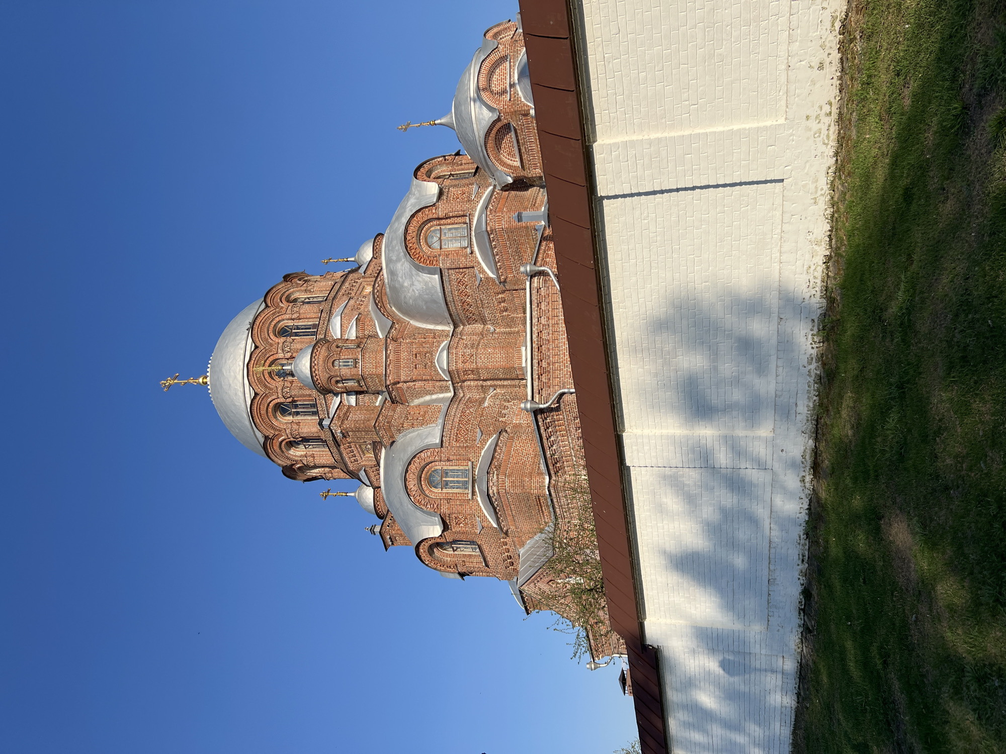 The image depicts the Cathedral of the Mother of God "Joy of All Who Sorrow" in Sviyazhsk, Russia, viewed from a low angle. The cathedral exhibits red brick construction with silver domes and golden crosses. A white brick wall runs alongside the cathedral, with green grass visible beyond the wall. The sky is a clear, deep blue. No people are visible. A shadow of a tree falls on the white brick wall.