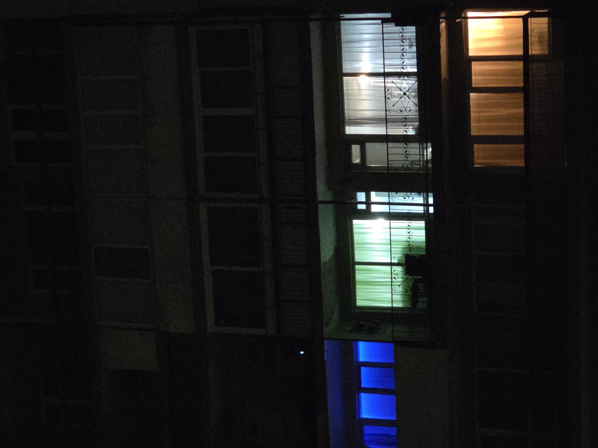 The image depicts the exterior of a building at night. Several windows are visible, illuminated by different colors. The window on the right emits an orange glow. The central window is lit in shades of white, green, and blue, with ironwork visible on the window. Other windows are dark and blend into the night. Location information (Pleven, Bulgaria) is outside the scope of observable detail.