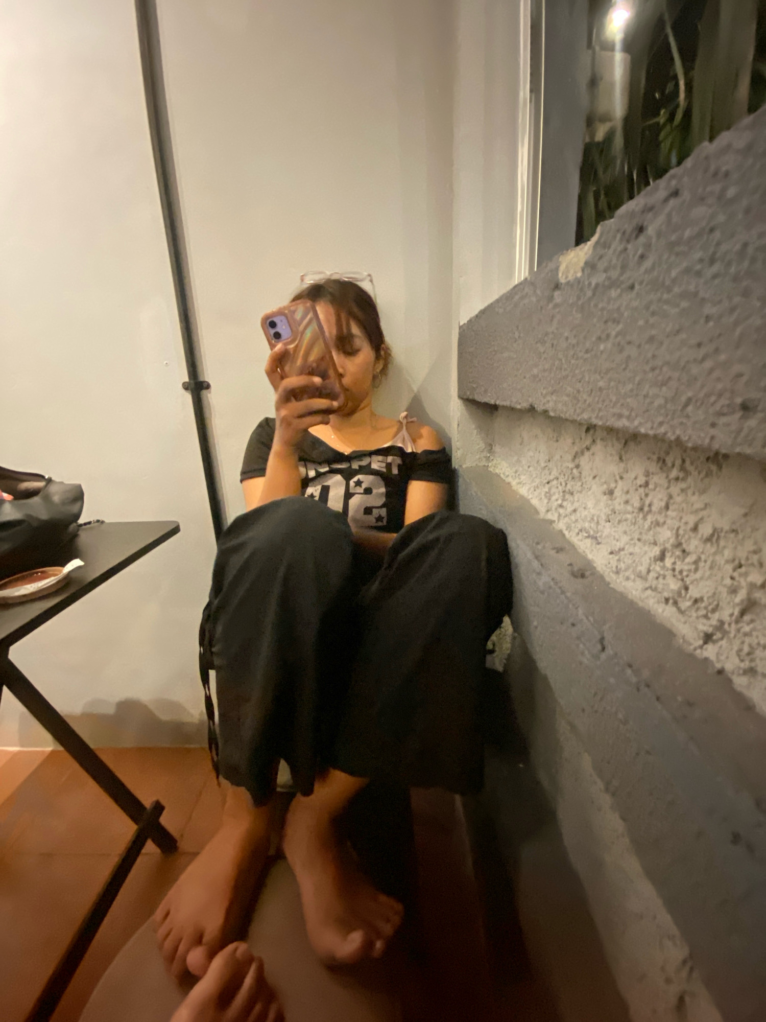 A young woman with glasses perched on her head is sitting on the floor in a corner, holding a smartphone and looking at its screen. She is wearing a black off-the-shoulder top with a white graphic and black wide-leg pants. Her bare feet are visible in front of her. To her left, a small black table with a black bag and a plate is situated. The scene appears to be outdoors, with a textured concrete wall in the background and some foliage visible beyond it. The lighting suggests it is evening or nighttime. This image is likely from Klurak, Indonesia, given the context.