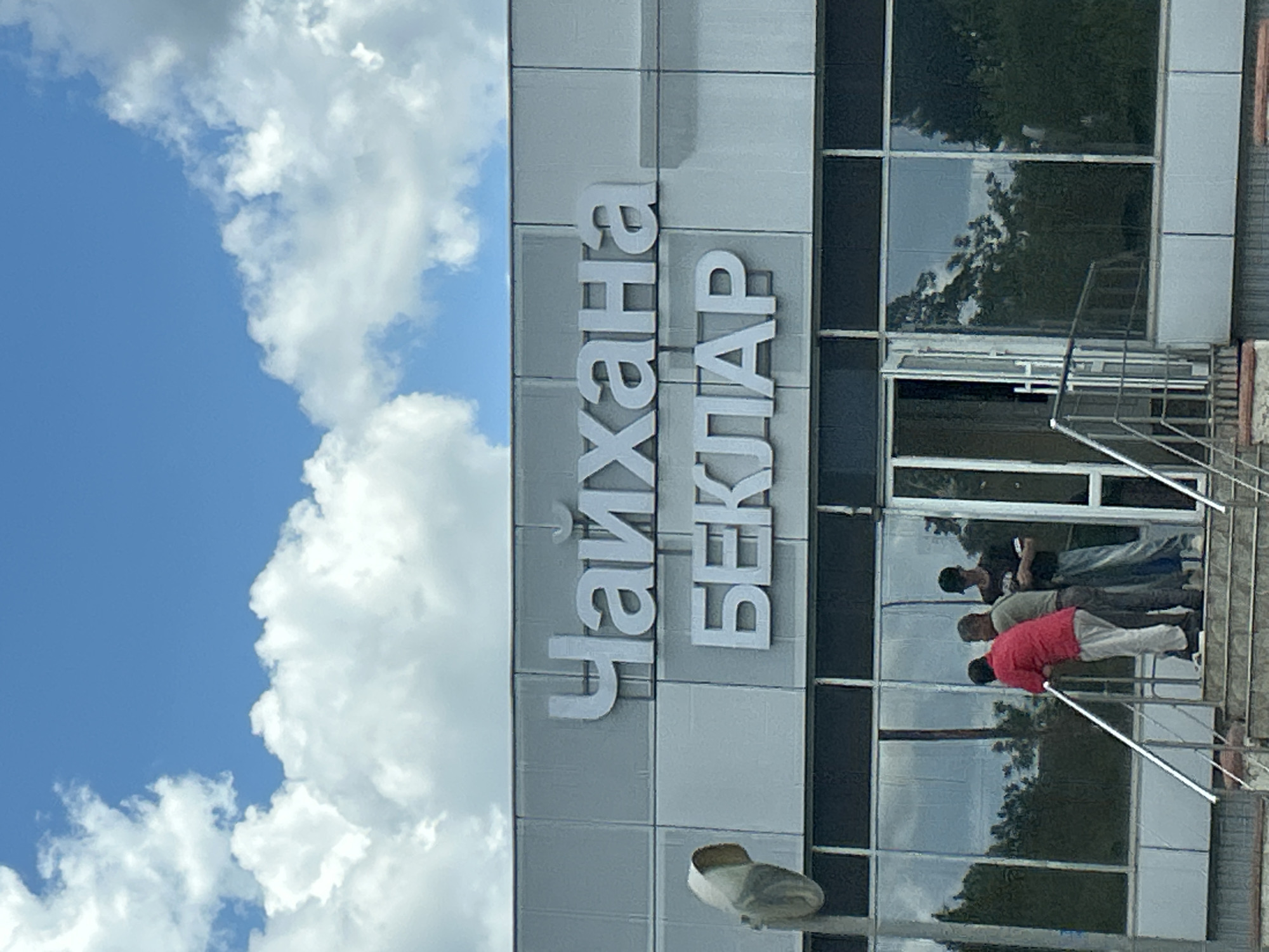 This image shows the exterior of a building with large glass windows and gray paneling. White, three-dimensional lettering is mounted vertically on the building's facade, spelling out "ЧАЙХАНА БЕКЛАР". The word "Чайхана" translates to "tea house" or "tea garden," and "Беклар" is likely a name or place.
In front of the building, there are several people, partially visible through the glass windows and on a set of outdoor stairs. The individuals appear to be wearing casual clothing. One person in the foreground on the stairs is wearing a red jacket. The reflections in the windows show trees and the sky.
The scene is outdoors, indicated by the bright blue sky and fluffy white clouds that dominate the left side of the image. The lighting suggests it is daytime, likely sunny. The architecture of the building, with its modern facade and glass panels, along with the presence of an outdoor staircase, hints at a commercial establishment, possibly a restaurant or cafe. The overall impression is one of a bright, possibly warm day in a developed area.