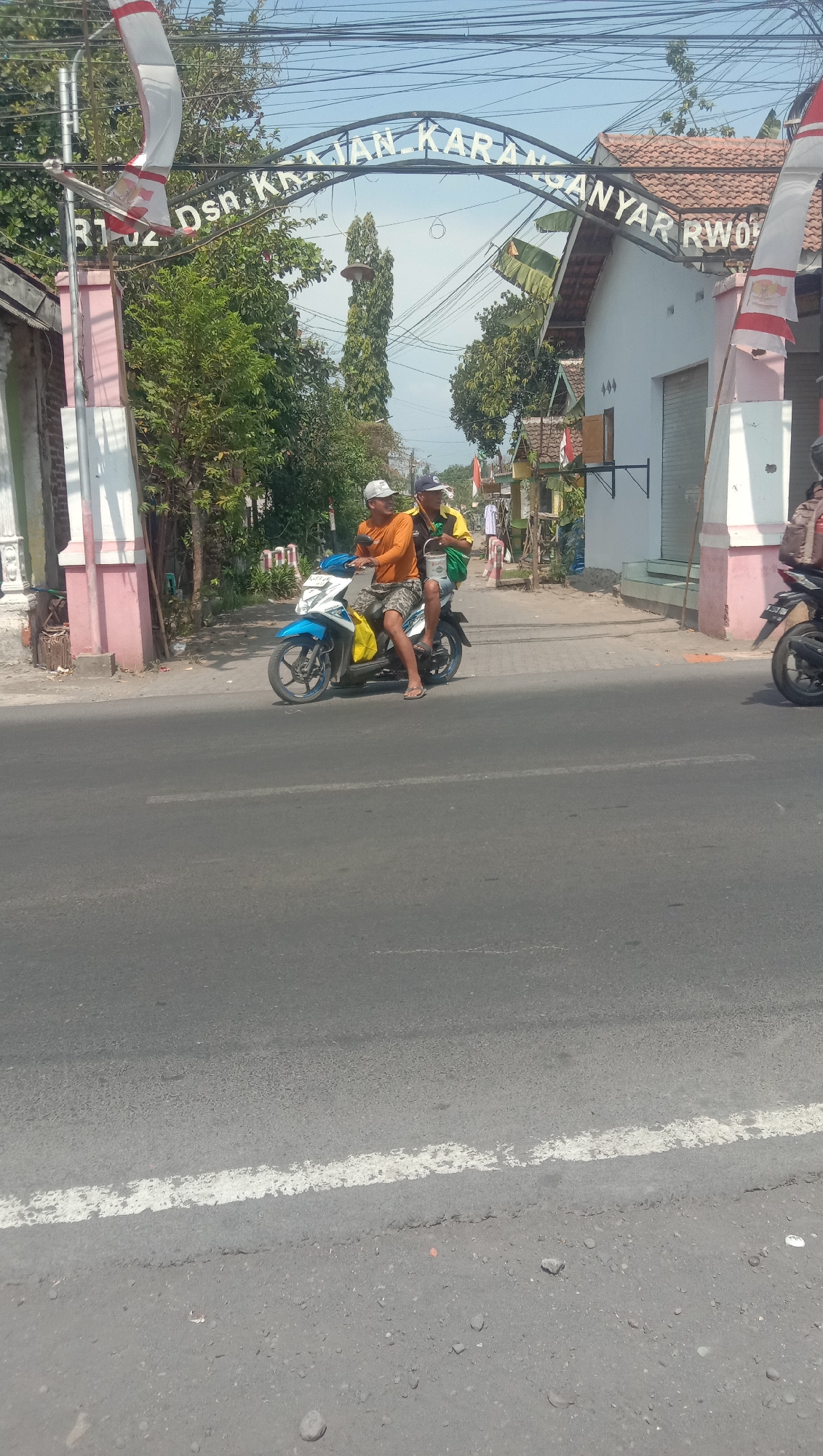Two men are riding on a light blue and white motorcycle on a paved road. The man in the front is wearing an orange shirt and patterned shorts, and the man behind him is wearing a yellow and black shirt and dark shorts. Both are wearing hats. The man in the back is holding a white bucket. They are passing under an archway with text on it that reads "RT 02 DSN. KRAJAN KARANG ANYAR RWO". The archway is decorated with Indonesian flags. The background shows buildings and trees, and the sky is clear and blue. The image appears to have been taken in Karang Anyar, Indonesia.