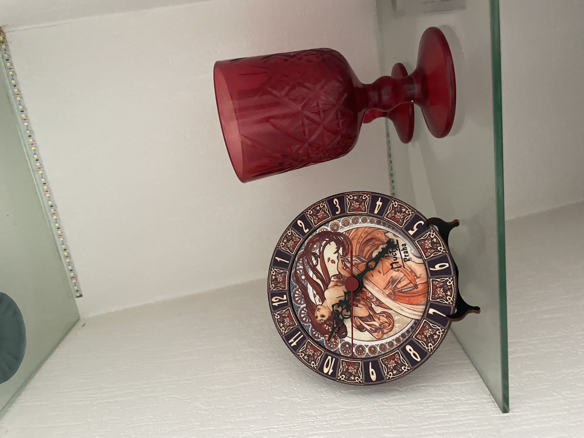 The image depicts a glass shelf with a red goblet and a clock. The clock features an Art Nouveau design with figures and the words "Mucha Praha." The surrounding environment appears to be an interior space with white walls and possible LED lighting along the edges of a structure. The image provides insufficient data to confidently determine that the location is in the city of Krasnoyarsk, Russia.