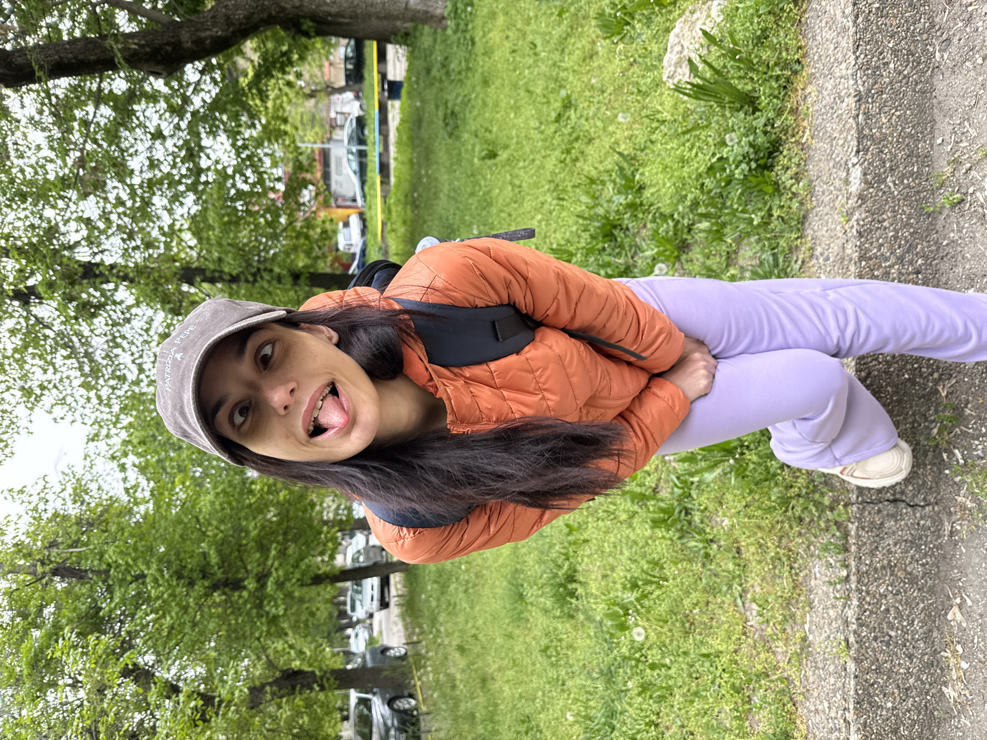 A young woman is photographed outside in Pleven, Bulgaria. She wears a grey baseball cap, an orange puffer jacket, and lavender sweatpants with white shoes. She is standing on a concrete sidewalk next to a grassy area with trees in the background. Her tongue is sticking out. Parked cars are visible in the background behind yellow construction tape.
