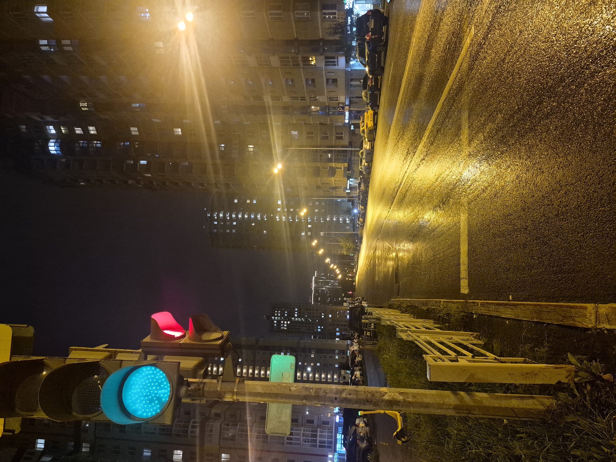 This image captures a nighttime street scene in Yekaterinburg, Russia. A traffic light in the foreground displays a cyan-blue light on its top signal and a red light on its middle signal. To the left of the traffic light, a yellow sign with green text is mounted on a pole, and a yellow electric scooter is parked on the grass behind the pole. A metal fence with yellow railings runs along the side of the road, separating the grass from the wet asphalt. The road itself reflects the light from street lamps and the windows of tall apartment buildings lining the right side of the street. Several cars are parked along the right side of the road. The buildings are illuminated by numerous lit windows, indicating occupancy. The overall atmosphere is that of a quiet, wet, urban night.