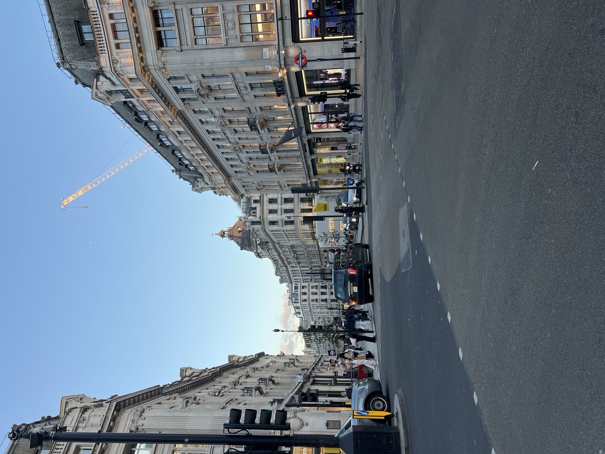 The image depicts a street intersection in a city, likely London given the presence of red double-decker buses and the iconic Underground logo. The architecture is grand, with ornate stone buildings lining the streets, featuring multiple stories, large windows, and decorative detailing. A construction crane is visible in the distance, suggesting ongoing development. Several vehicles, including cars and a black taxi, are present on the road. Pedestrians are crossing the street and walking on the sidewalks. The sky is a clear blue with a hint of twilight. The scene is a typical urban streetscape with a blend of commerce, transportation, and pedestrian activity.