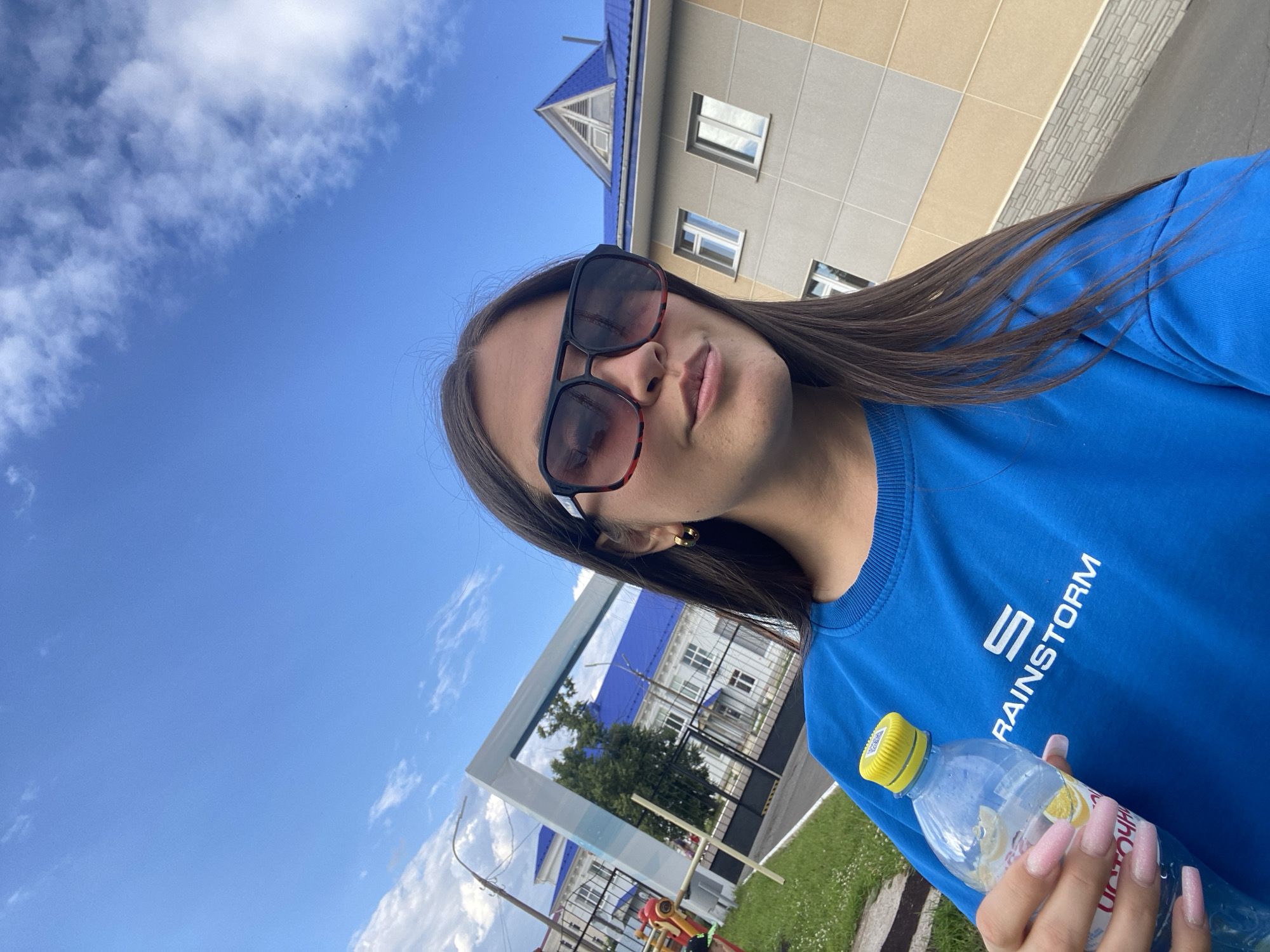 A young woman is taking a selfie outdoors on a sunny day. She has long, dark hair, is wearing large, dark sunglasses and a bright blue sweatshirt with white text that reads "S RAINSTORM". She is holding a clear plastic water bottle with a yellow cap in her right hand, which has long, light pink, sparkly fingernails. In the background, there is a light-colored building with a blue roof and windows. To the left, the sky is a clear blue with scattered white clouds. The scene appears to be in Chaikovsky, Russia. The overall mood is casual and relaxed.