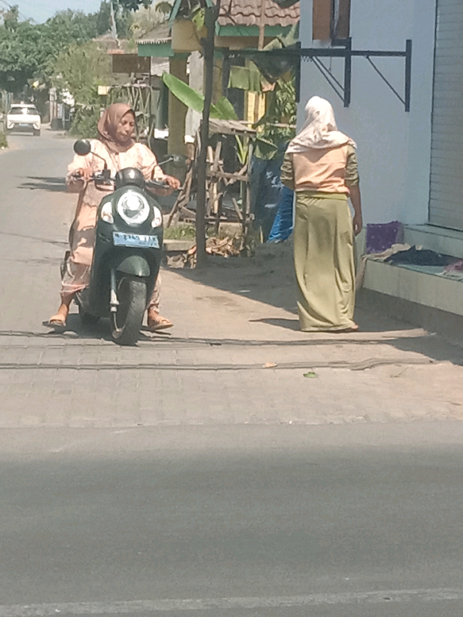 In the city of Karang Anyar, Indonesia, two women are visible on a street. One woman, wearing a peach-colored tunic and a light beige hijab, is riding a dark-colored scooter. She is positioned on the left side of the frame and is looking forward. The scooter has a visible license plate. Another woman, wearing a light orange long-sleeved shirt and a long, olive-green skirt, stands on the right side of the image, facing away from the camera. She is looking towards the building on the right. The environment appears to be a semi-urban or rural area with a road in the foreground and buildings and vegetation in the background. The lighting suggests it is daytime.